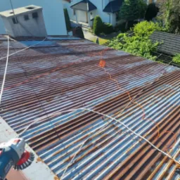 Przygotowanie zardzewiałej blachy falistej na dachu do dalszych prac, szlifierka kątowa z drucianą szczotką w użyciu, widoczny przewód elektryczny i lina zabezpieczająca.