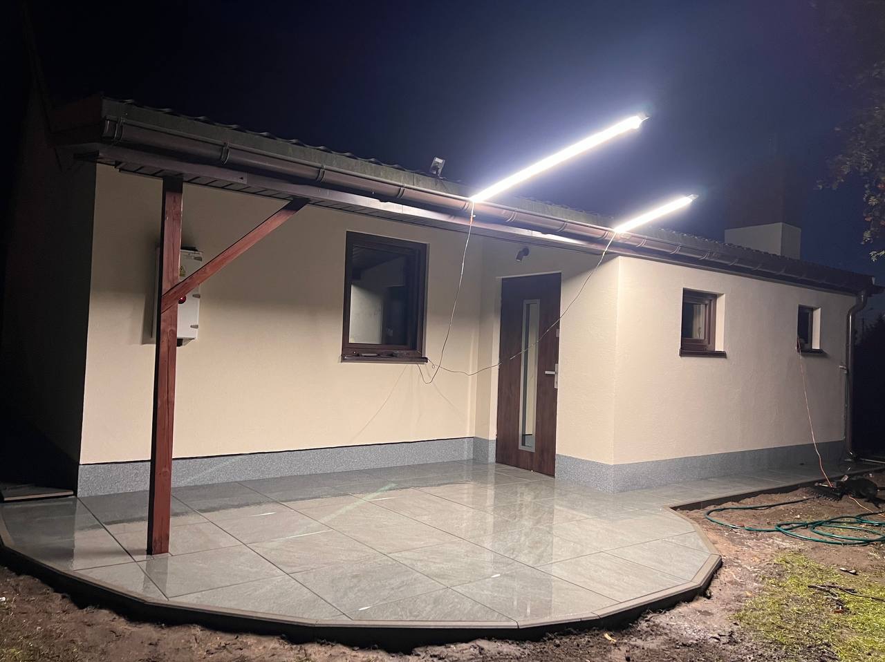 Elewacja domu jednorodzinnego w nocy, oświetlona dwoma lampami LED, z nową, szarą, ceramiczną podłogą tarasu i świeżo pomalowaną fasadą w kolorze beżowym.
