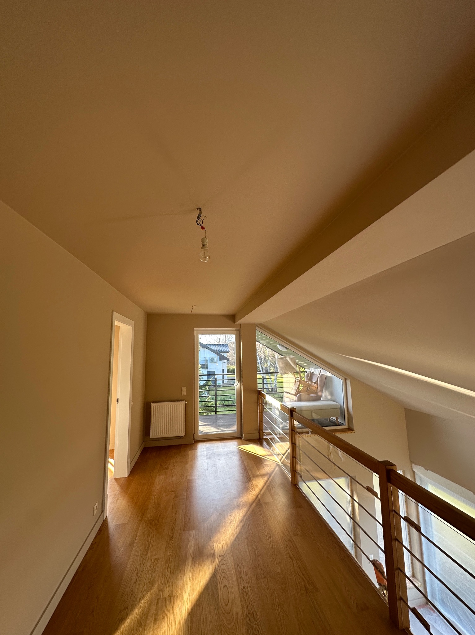 Poddasze po malowaniu: beżowe ściany, drewniana podłoga, balkon z widokiem na zieleń. Widoczne gniazdko bez lampy, grzejnik i drewniane barierki. Słoneczne światło.