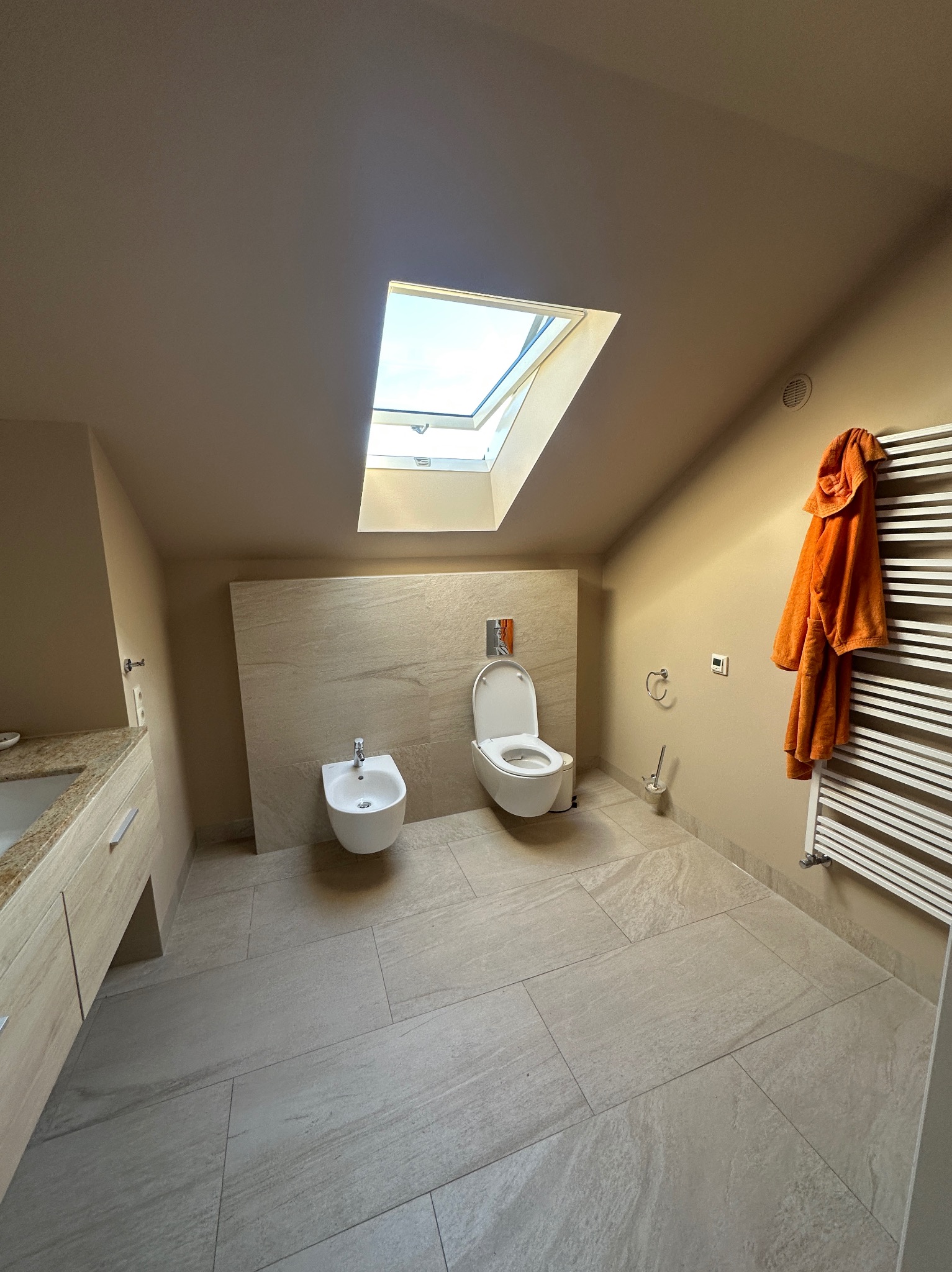 Jasna łazienka na poddaszu z oknem dachowym, beżowymi płytkami i białymi sanitariatami. Pomarańczowy szlafrok na grzejniku dodaje koloru. Widok z góry.