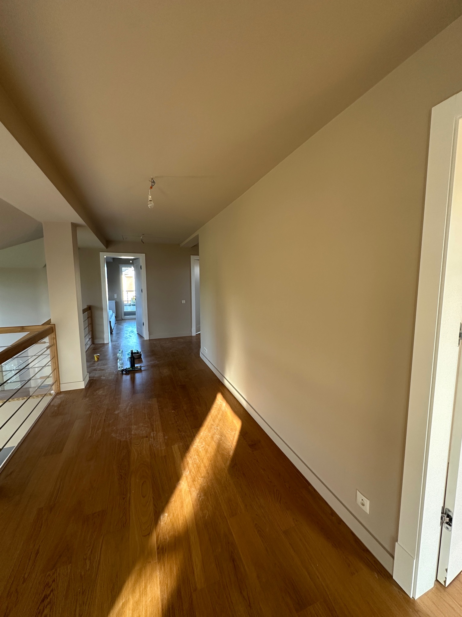 Jasne wnętrze po malowaniu: beżowe ściany, drewniana podłoga z refleksami słońca. Widoczne narzędzia malarskie w oddali, białe listwy przypodłogowe i ościeżnice.