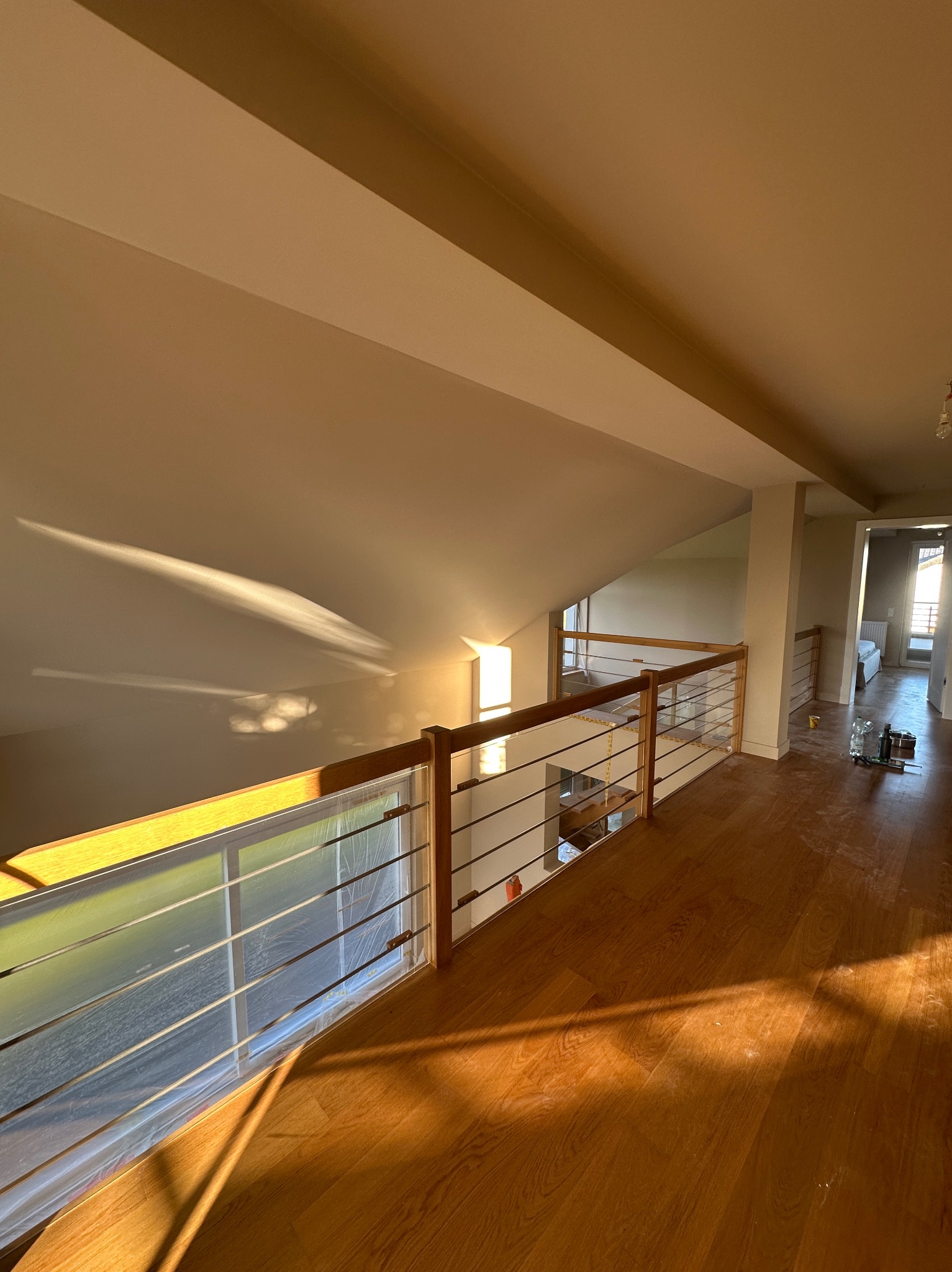 Jasne wnętrze poddasza z drewnianą podłogą i balustradą, ściany świeżo pomalowane na kremowo, okna zabezpieczone folią malarską, wpadające promienie słoneczne.