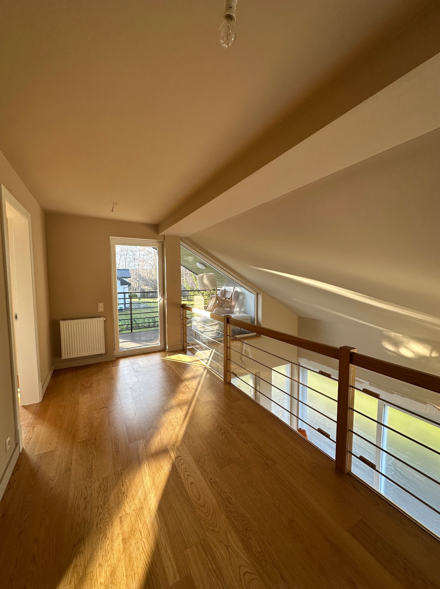 Jasne, przestronne poddasze z drewnianą podłogą i balustradą. Ściany w odcieniach beżu, wpadające promienie słoneczne. Widok na zewnątrz przez balkonowe drzwi.