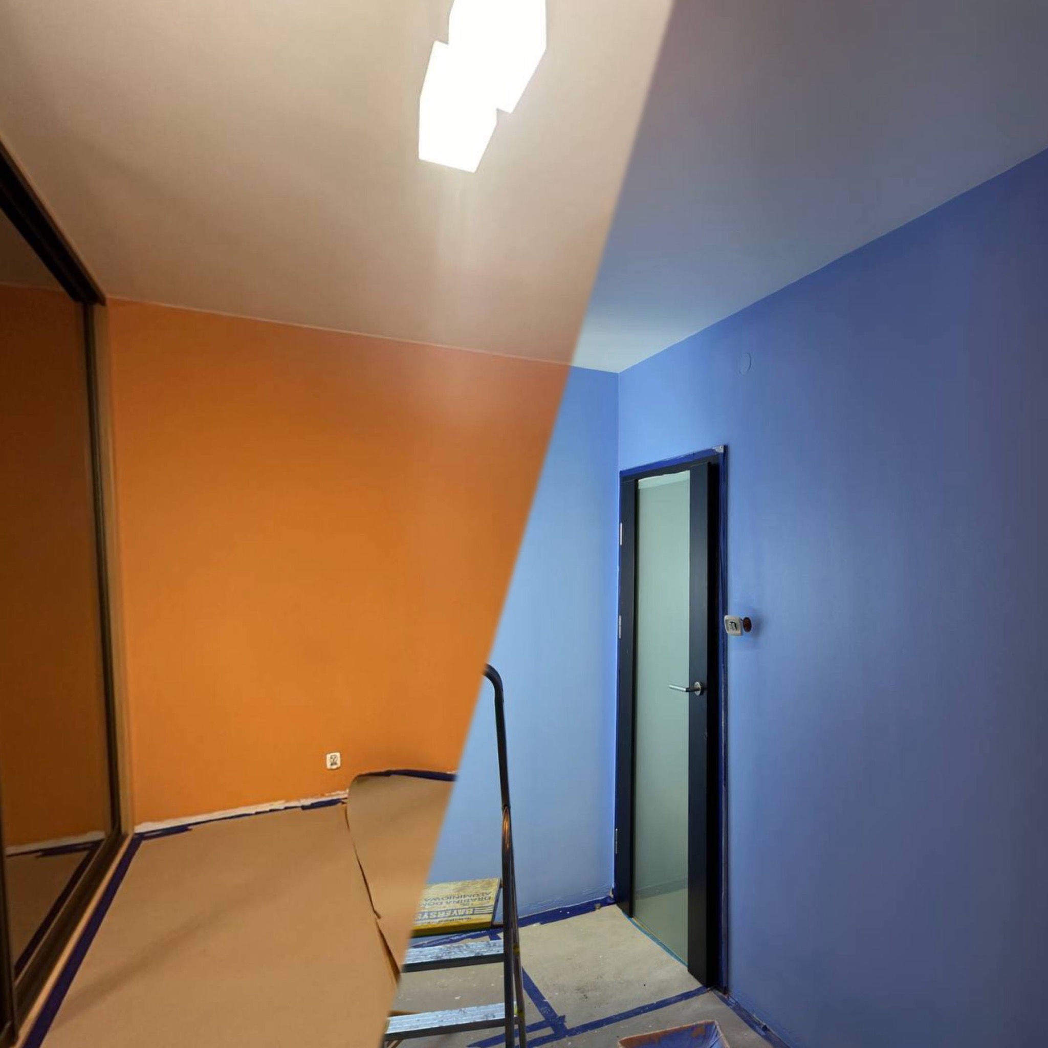 Pokój po częściowym malowaniu: ściana pomarańczowa i niebieska, zabezpieczona podłoga, drabina. Widoczny efekt zmiany koloru w trakcie prac.