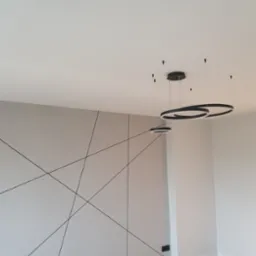 Minimalistyczne wnętrze z szarą ścianą zdobioną czarnymi, geometrycznymi liniami, nowoczesnymi lampami wiszącymi w kształcie pierścieni i jasną podłogą.