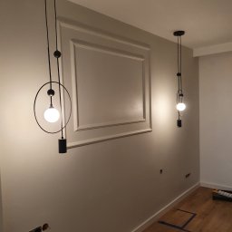 ŁUKASZ KALETA ARTEMIL - Szare wnętrze z dwiema nowoczesnymi lampami wiszącymi z okrągłym kloszem i dekoracyjną ramą na ścianie, widoczne niedokończone gniazdka elektryczne i listwy przypodłogowe.