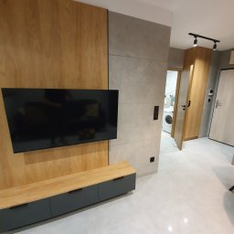 ŁUKASZ KALETA ARTEMIL - Nowoczesny salon z drewnianą ścianą, telewizorem, szafką RTV z szufladami i widokiem na łazienkę z pralką w tle.