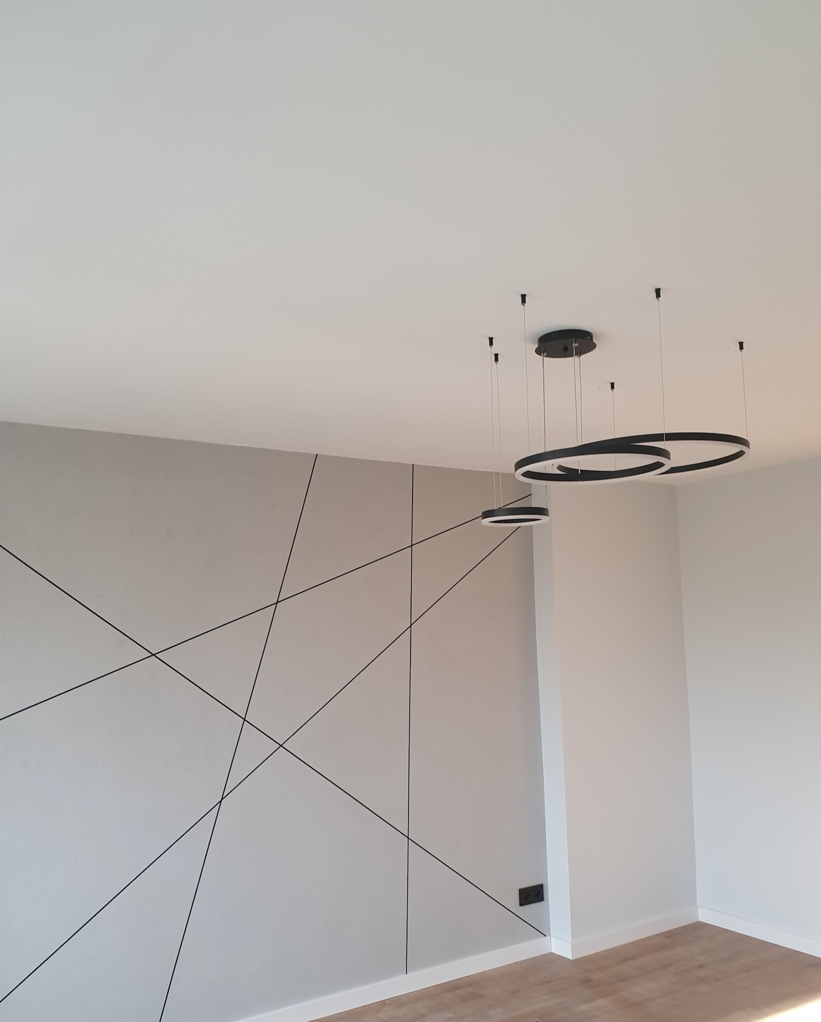 Minimalistyczne wnętrze z szarą ścianą zdobioną czarnymi, geometrycznymi liniami, nowoczesnymi lampami wiszącymi w kształcie pierścieni i jasną podłogą.