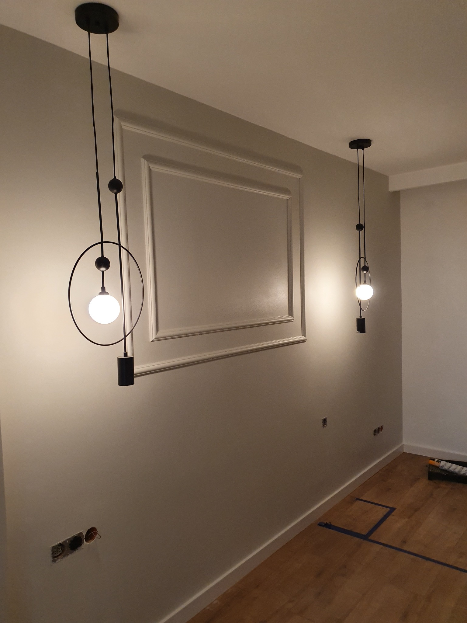 Szare wnętrze z dwiema nowoczesnymi lampami wiszącymi z okrągłym kloszem i dekoracyjną ramą na ścianie, widoczne niedokończone gniazdka elektryczne i listwy przypodłogowe.