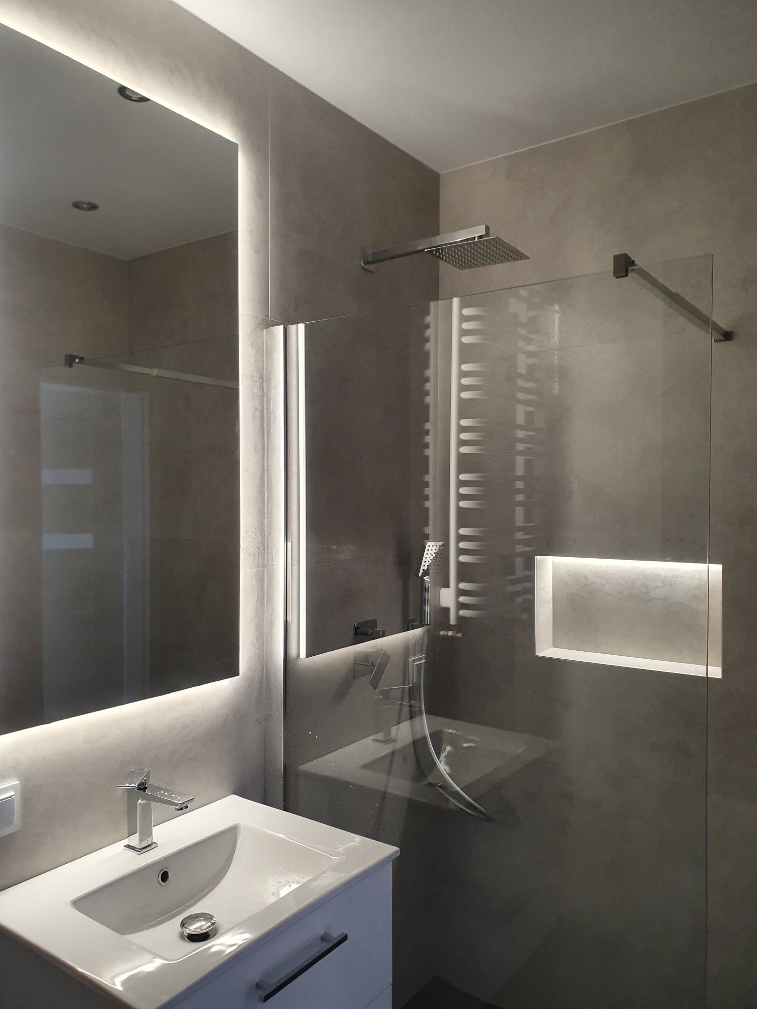 Minimalistyczna łazienka z kabiną prysznicową typu walk-in, podświetlanym lustrem i wnęką, wykończona w odcieniach szarości, z chromowaną armaturą.