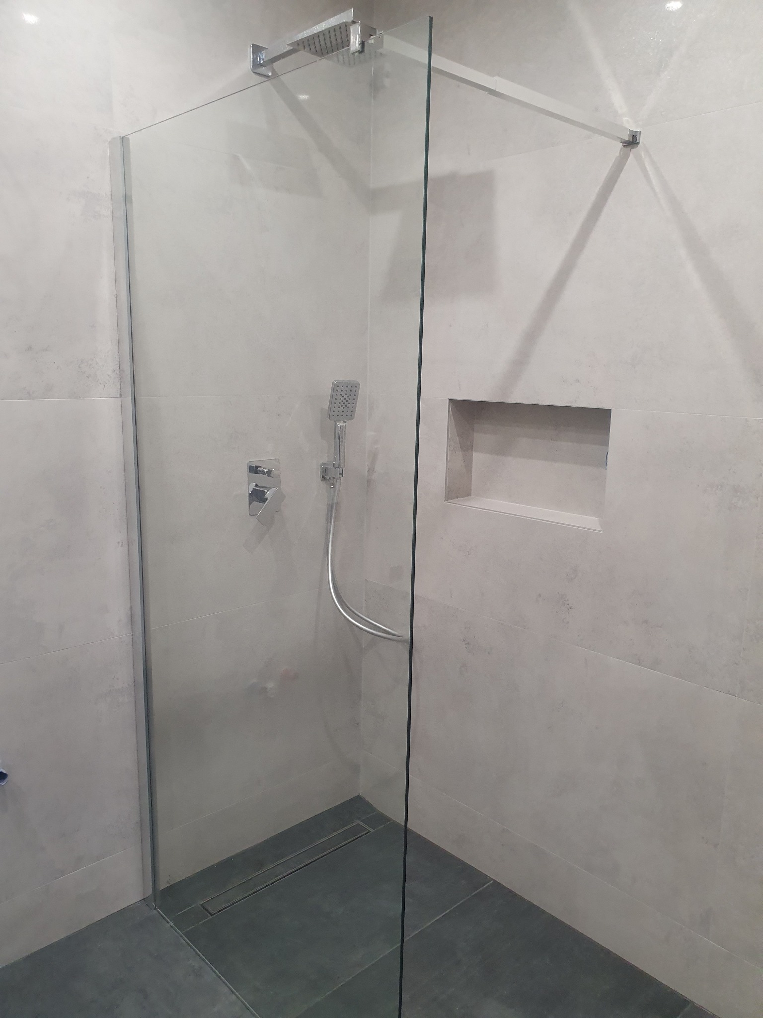 Nowoczesna kabina prysznicowa typu walk-in z odpływem liniowym, wykończona płytkami w odcieniach szarości, z wnęką w ścianie na kosmetyki.