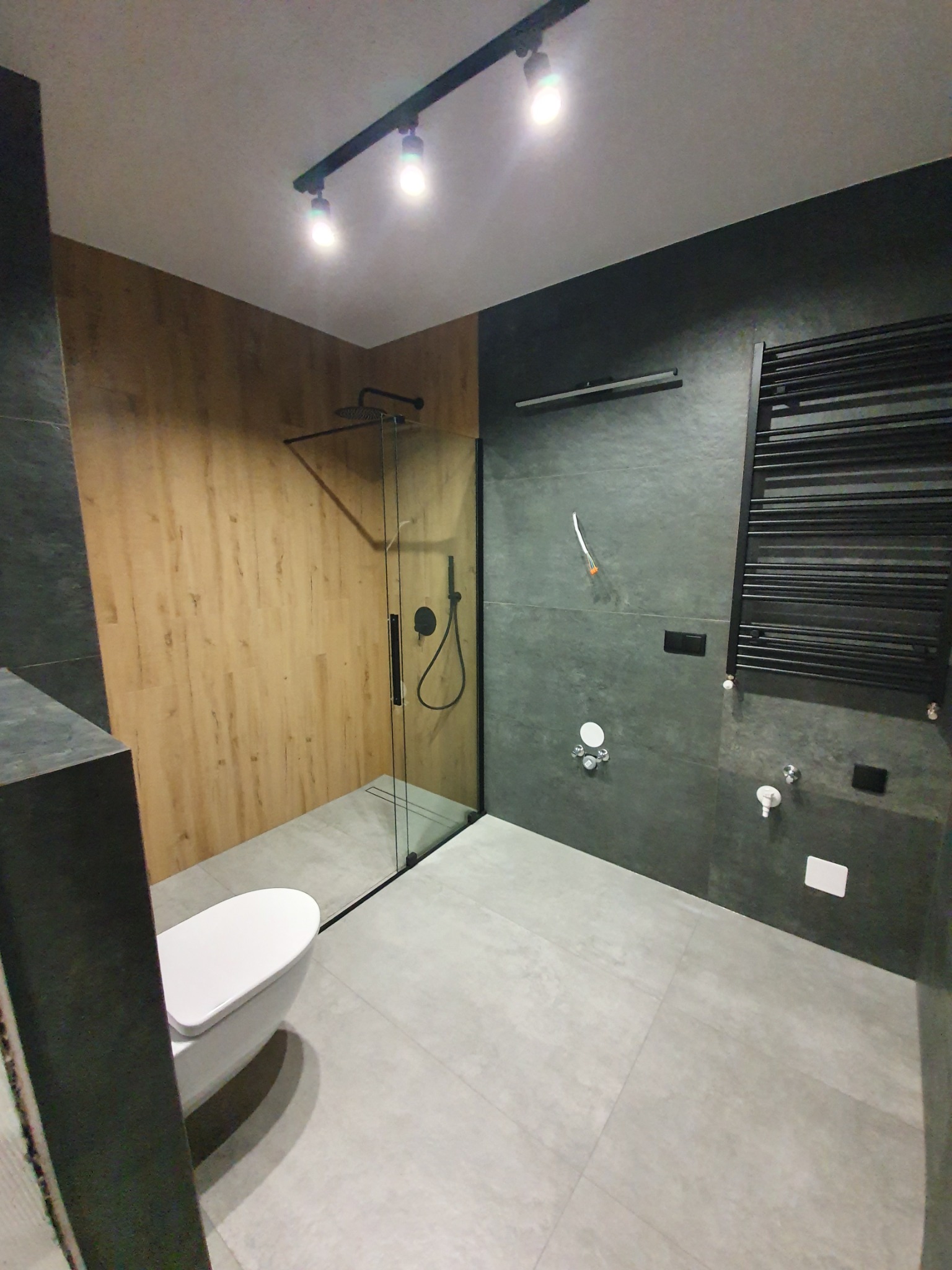 Nowoczesna łazienka z kabiną prysznicową typu walk-in, drewnopodobną okładziną ścienną, grafitowymi płytkami i grzejnikiem drabinkowym w czarnym kolorze, oświetlona reflektorami sufitowymi.