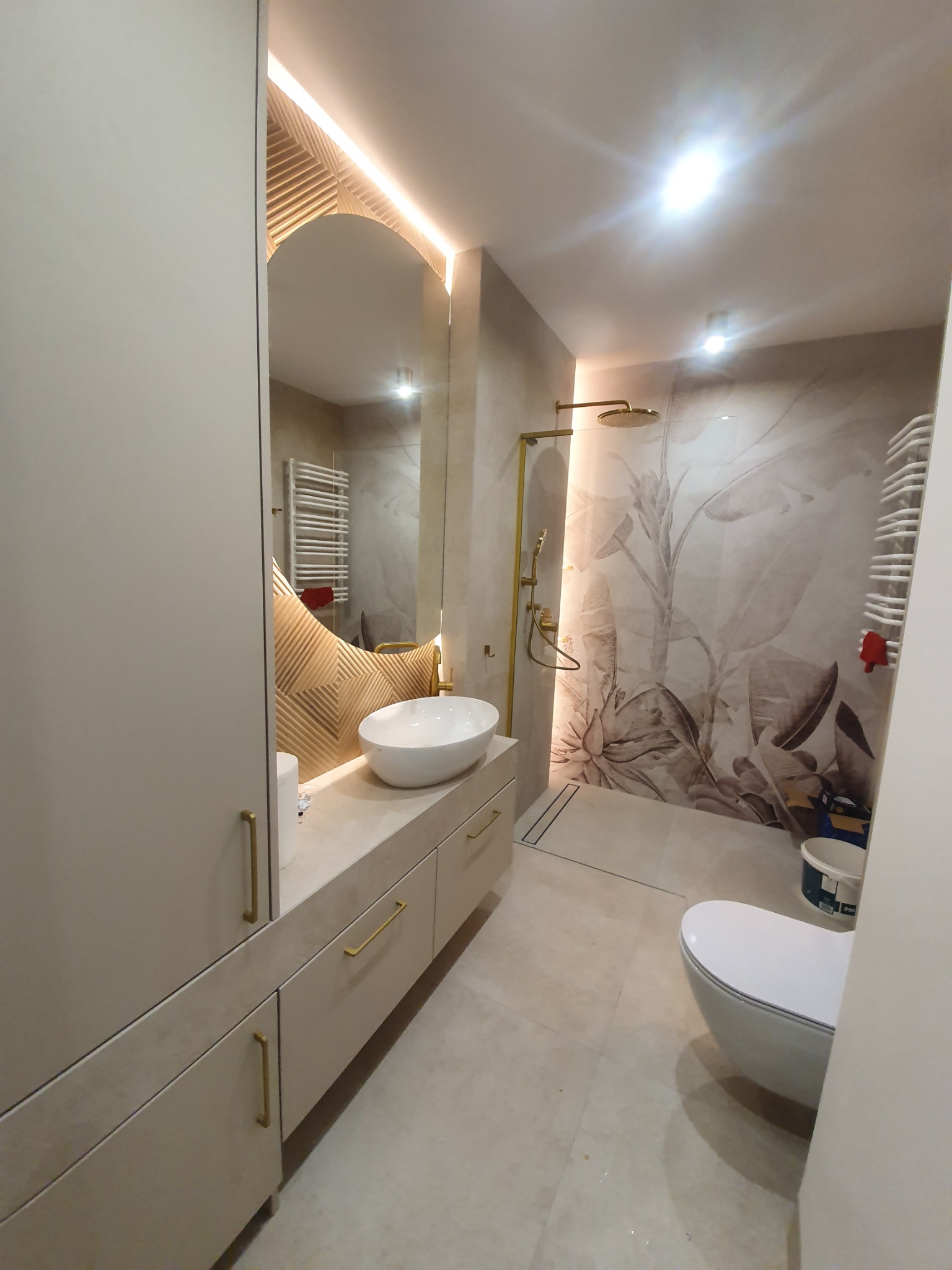 Nowoczesna łazienka z beżowymi szafkami, umywalką nablatową i lustrem z podświetleniem LED, kabina prysznicowa typu walk-in z deszczownicą i wzorzystą ścianą w motywy roślinne.