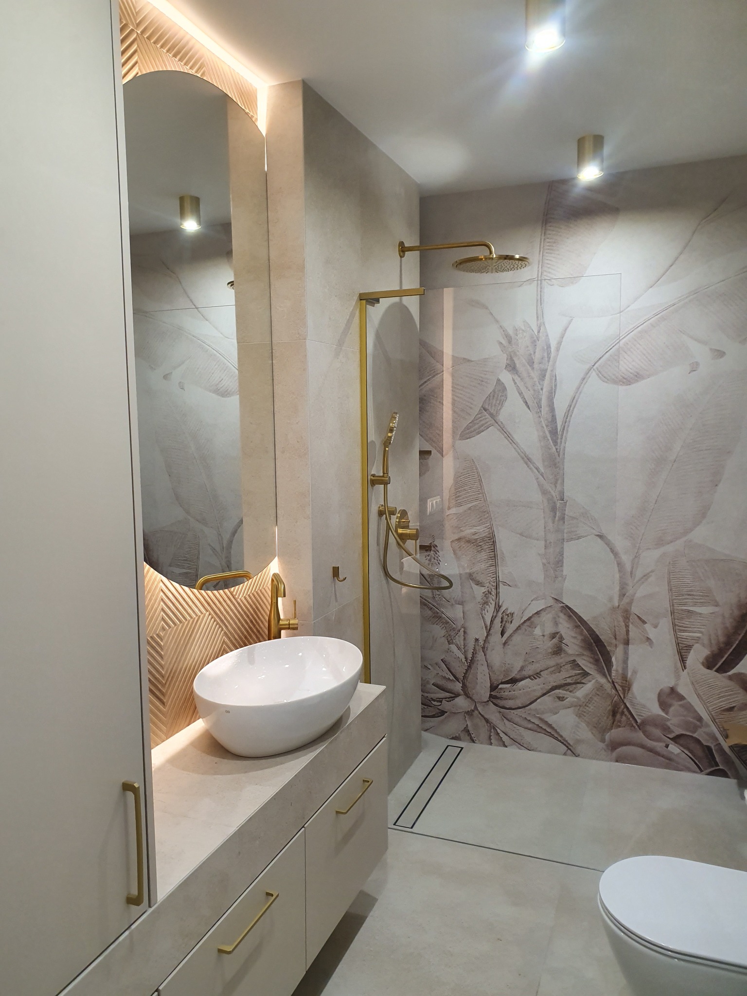 Nowoczesna łazienka z owalną umywalką nablatową, złotą armaturą i kabiną prysznicową typu walk-in z motywem roślinnym na ścianie, podświetlane lustro w kształcie łuku.