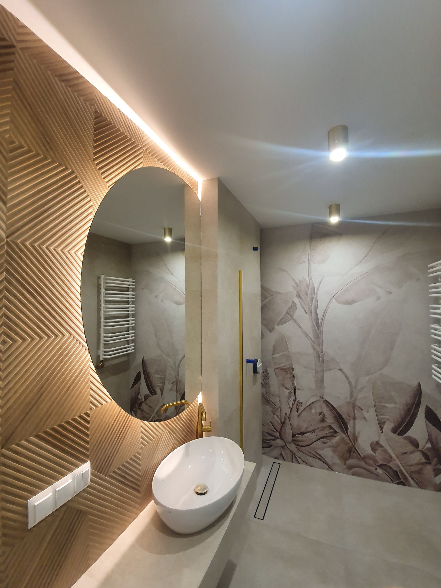 Nowoczesna łazienka z owalną umywalką nablatową, złotą armaturą, okrągłym lustrem podświetlonym LED, drewnianą ścianą z geometrycznym wzorem i tapetą w motywy roślinne.