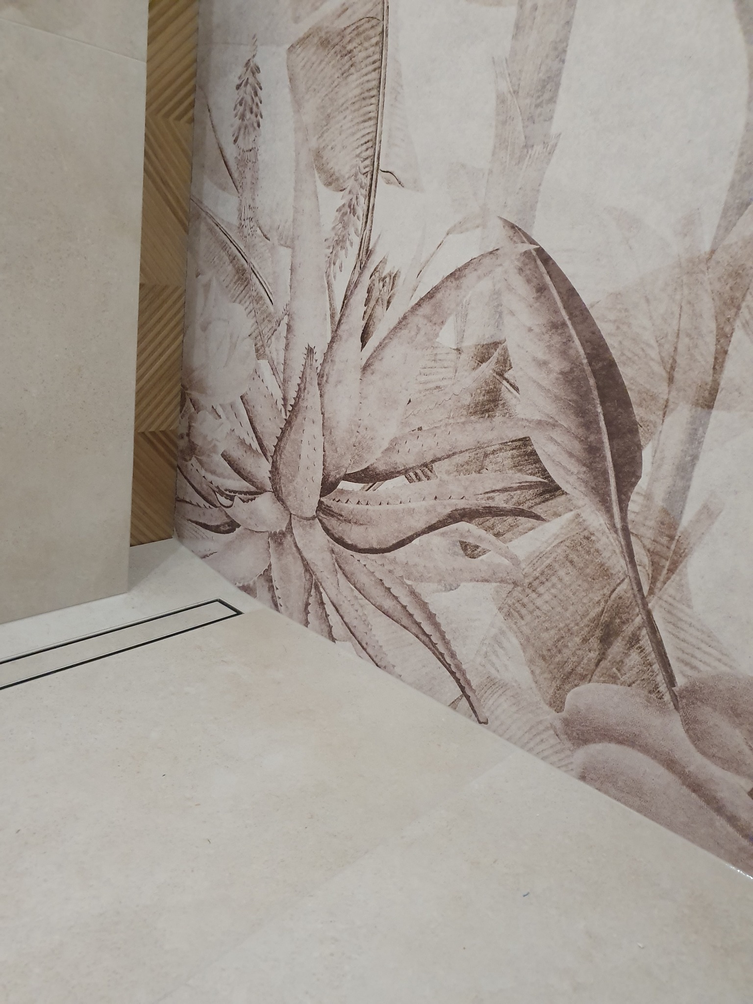 Beżowe płytki podłogowe i ścienne z motywem roślinnym w łazience, widoczny odpływ liniowy i dekoracyjna listwa.