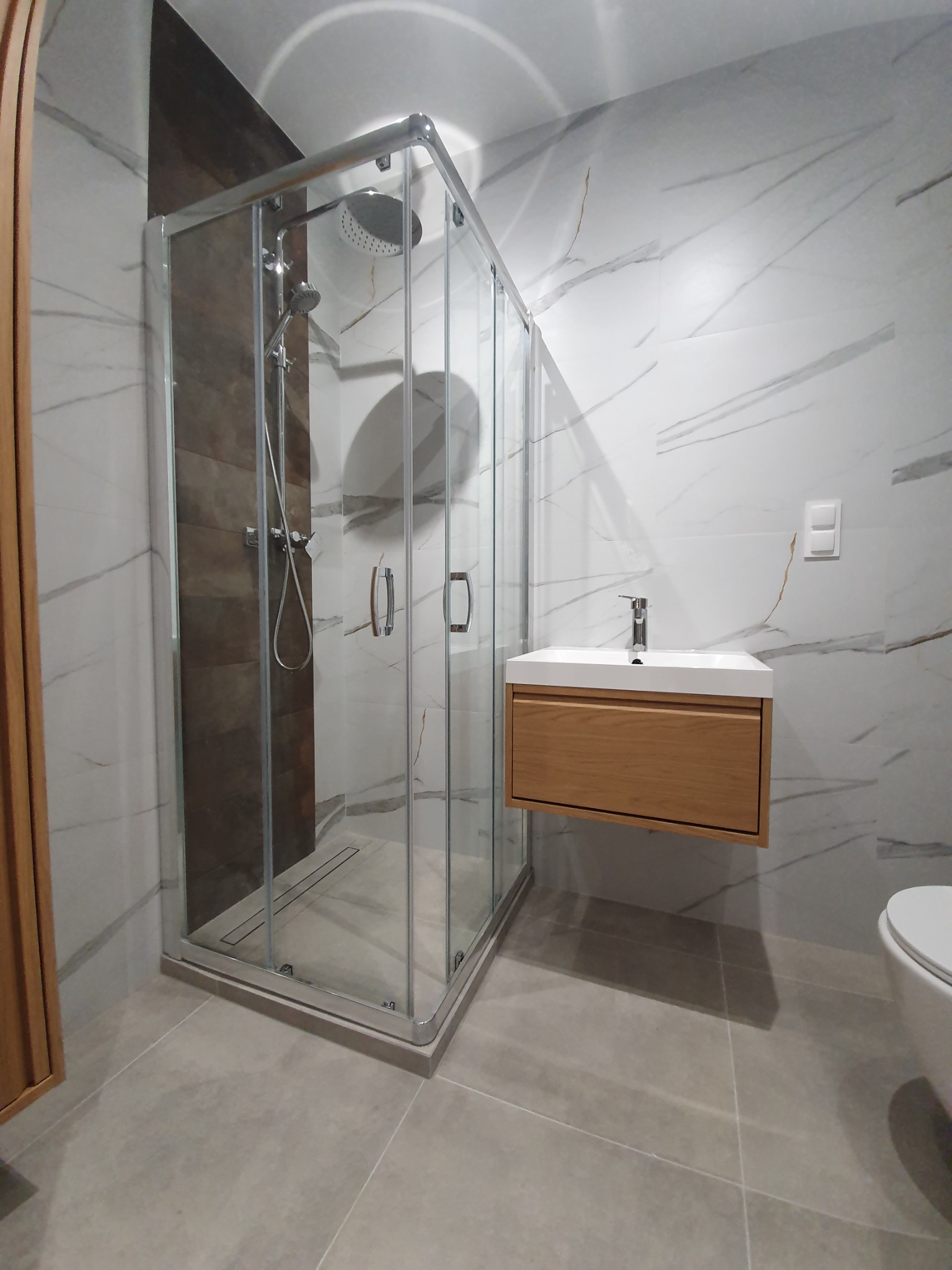 Nowoczesna łazienka w Kazimierzy Wielkiej z kabiną prysznicową ze szklanymi drzwiami, umywalką z drewnianą szafką i marmurowymi płytkami na ścianach.