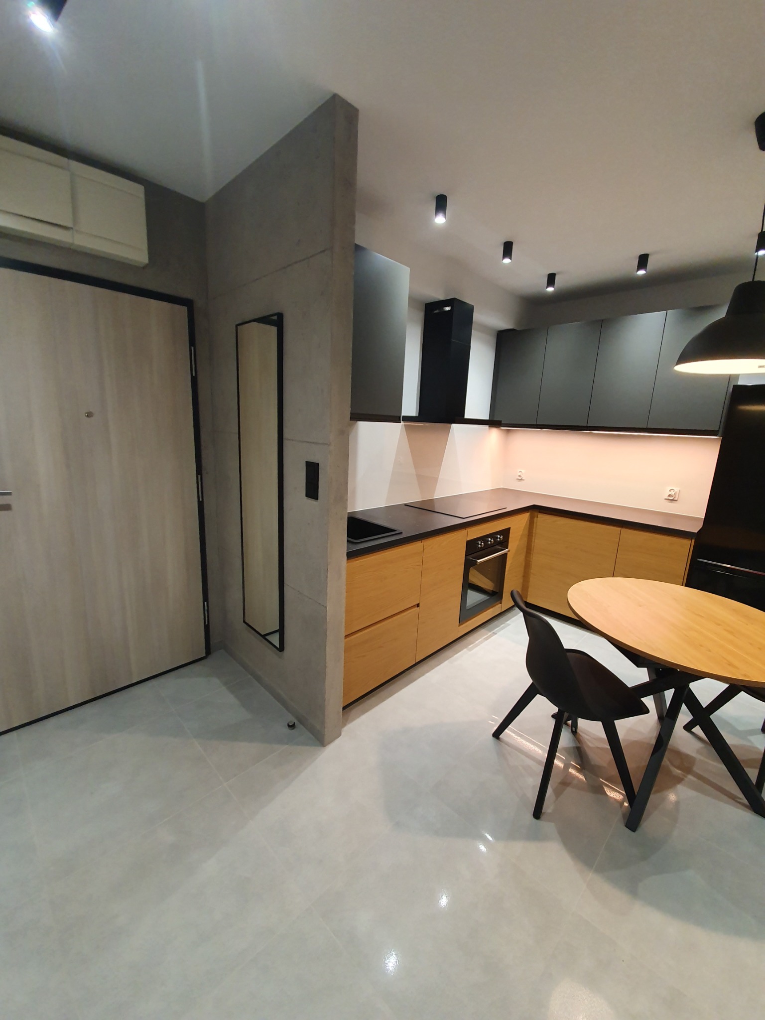 Nowoczesna kuchnia z drewnianymi frontami szafek, czarnymi górnymi szafkami i okrągłym stołem z czarnymi krzesłami, oświetlenie punktowe na suficie, minimalistyczne lustro na ścianie.