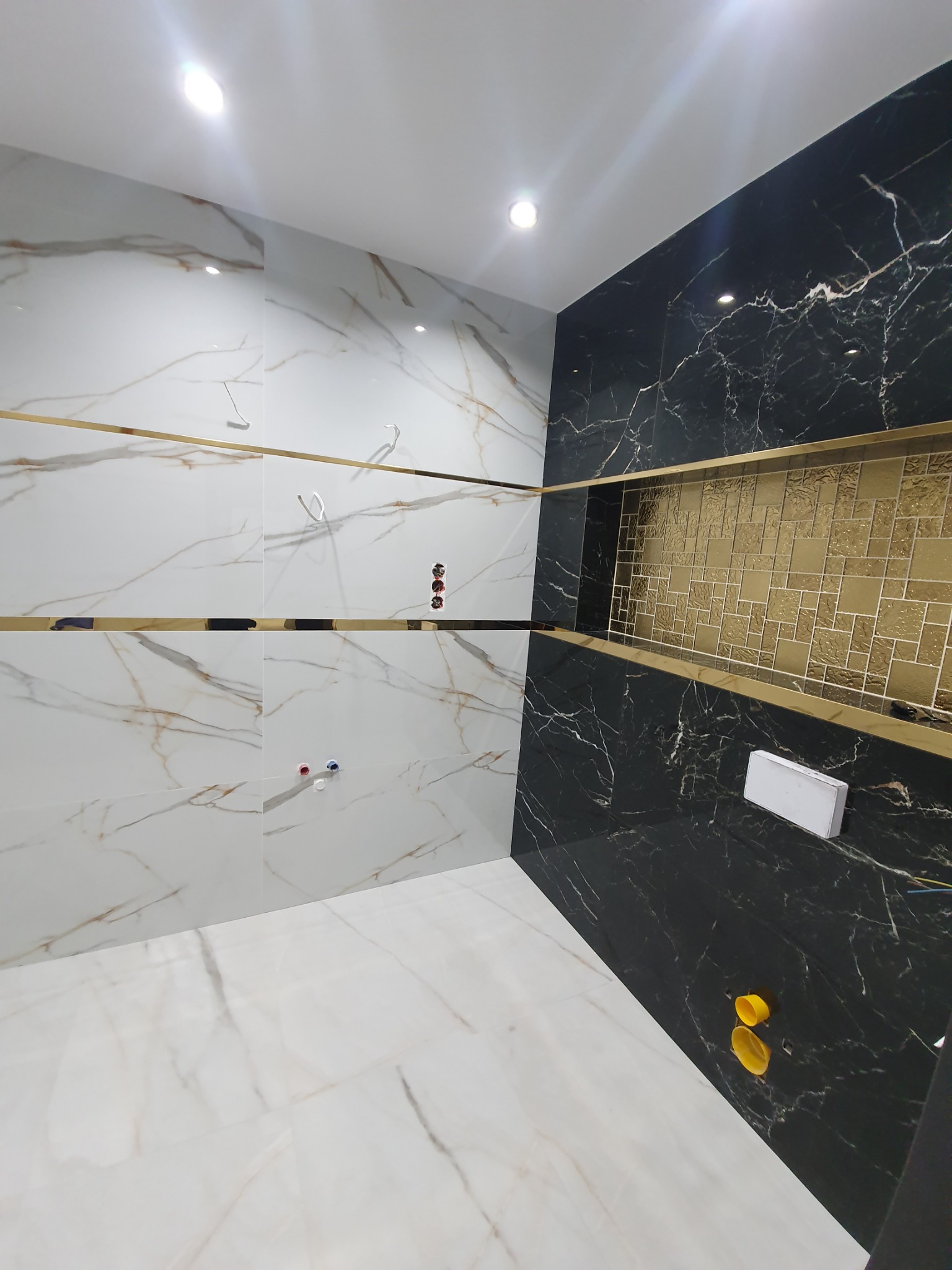 Nowoczesna łazienka w trakcie wykańczania, z białymi i czarnymi marmurowymi płytkami, złotymi listwami dekoracyjnymi, wnęką z mozaiką i wystającymi przewodami instalacji.