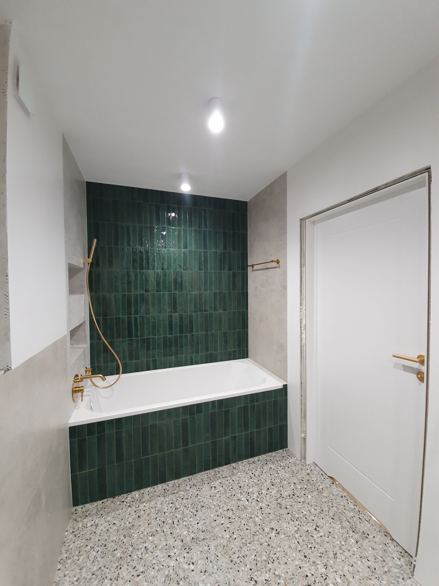 Wykończona łazienka z wanną obudowaną pionowymi, ciemnozielonymi płytkami, złotą armaturą i podłogą z lastryko.