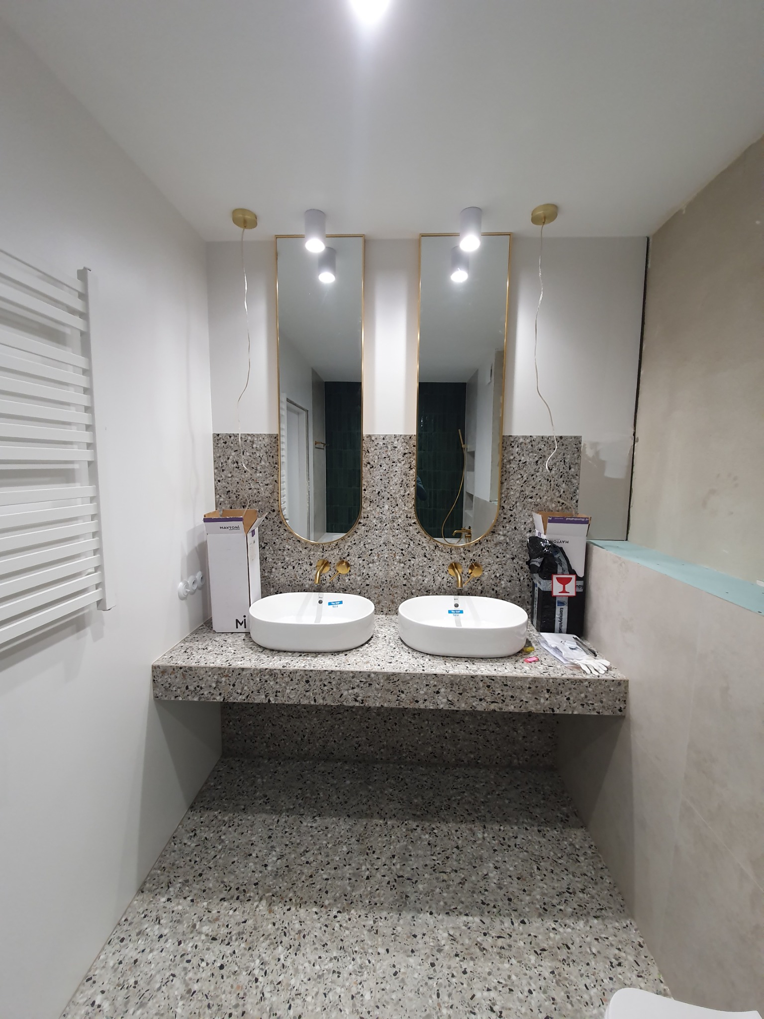 Dwie umywalki nablatowe w łazience wykończonej terazzo, z owalnymi lustrami w złotych ramach i białym grzejnikiem drabinkowym, podczas prac wykończeniowych.