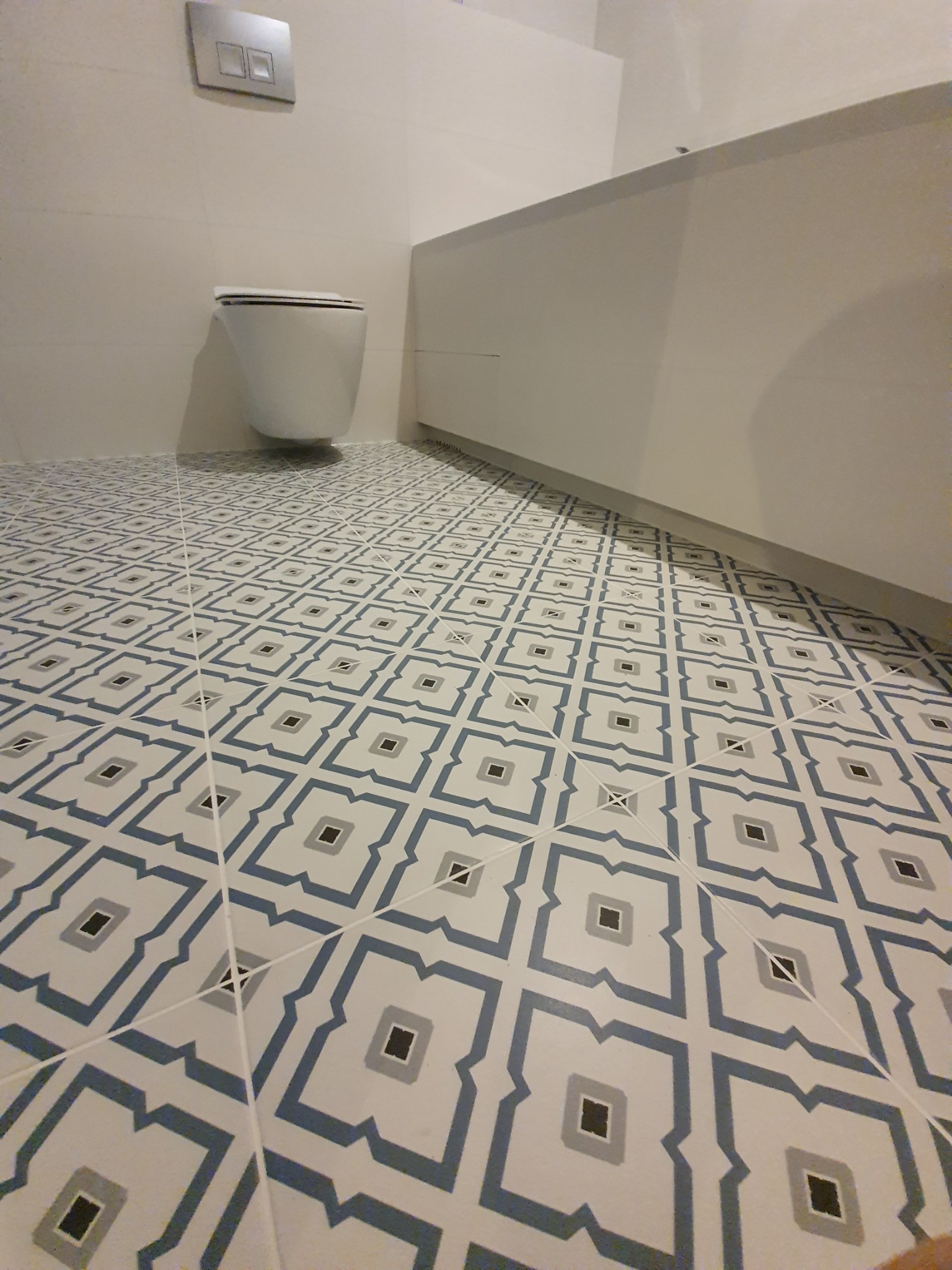 Wykończona łazienka z podwieszaną toaletą i wzorzystą, geometryczną podłogą w odcieniach bieli, szarości i błękitu.