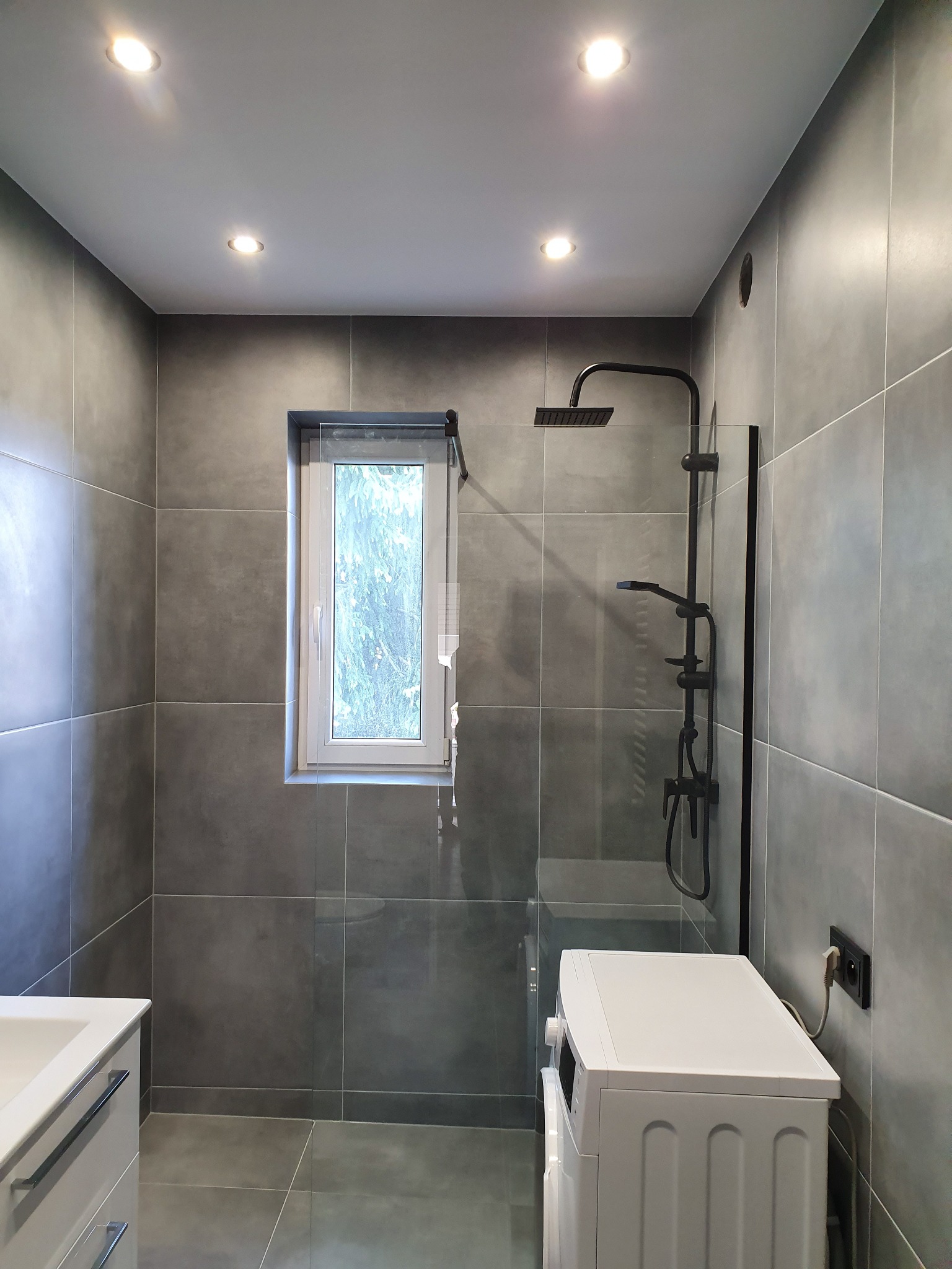 Nowoczesna łazienka z szarymi płytkami, czarną armaturą prysznicową, białą pralką i umywalką z szafką, oświetlona punktowo