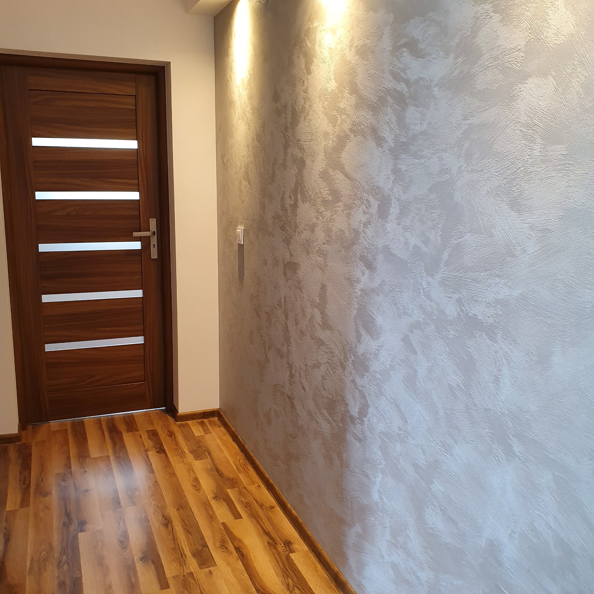 Ściana z dekoracyjnym, srebrzystym tynkiem strukturalnym, drewniana podłoga i nowoczesne drzwi z poziomymi przeszkleniami.