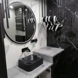 Nowoczesna łazienka z czarną umywalką nablatową, okrągłym lustrem z podświetleniem LED, czarno-białymi ręcznikami i marmurowymi płytkami na ścianach, z widoczną pralką pod blatem.