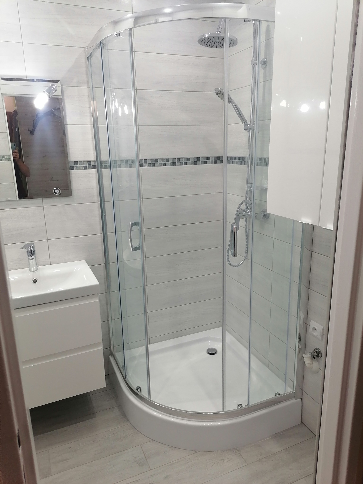 Nowoczesna łazienka z kabiną prysznicową narożną, umywalką z szafką w kolorze białym, szarymi płytkami imitującymi drewno na podłodze i mozaiką na ścianie.