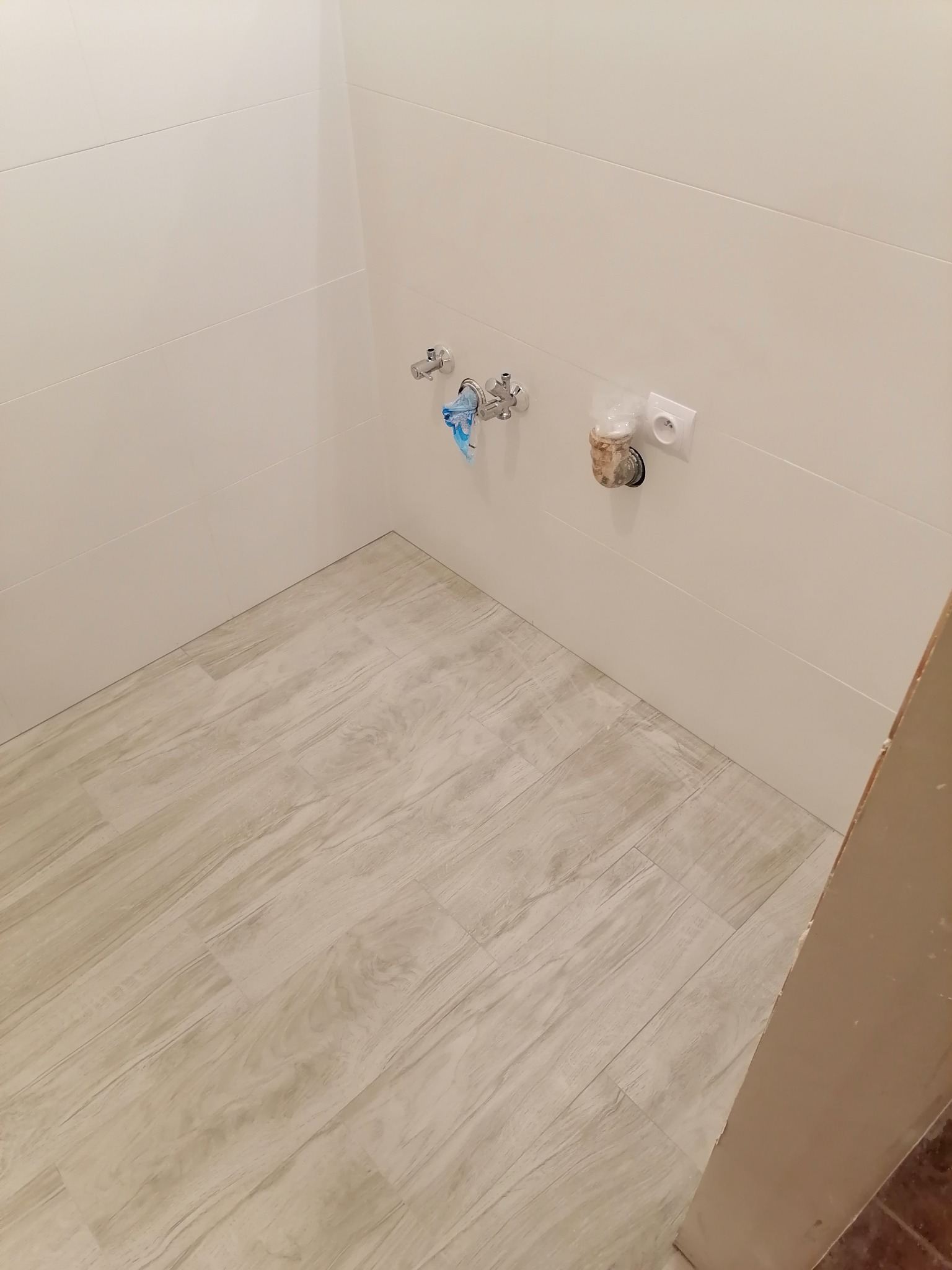 Świeżo wykończona łazienka z beżowymi płytkami imitującymi drewno na podłodze, białe płytki na ścianach, chromowana bateria umywalkowa z niebieską ściereczką i gniazdko elektryczne z wystającymi...