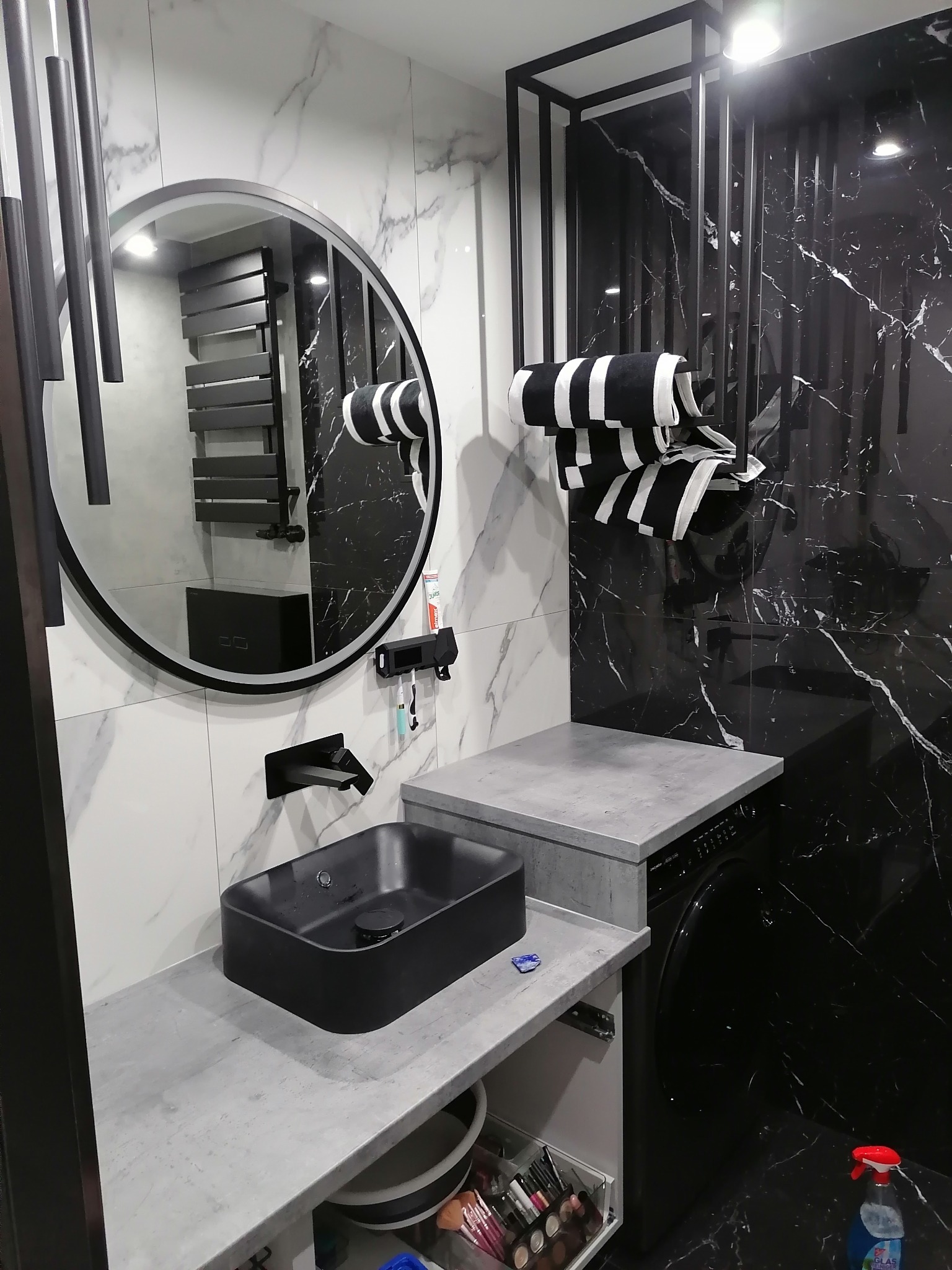 Nowoczesna łazienka z czarną umywalką nablatową, okrągłym lustrem z podświetleniem LED, czarno-białymi ręcznikami i marmurowymi płytkami na ścianach, z widoczną pralką pod blatem.