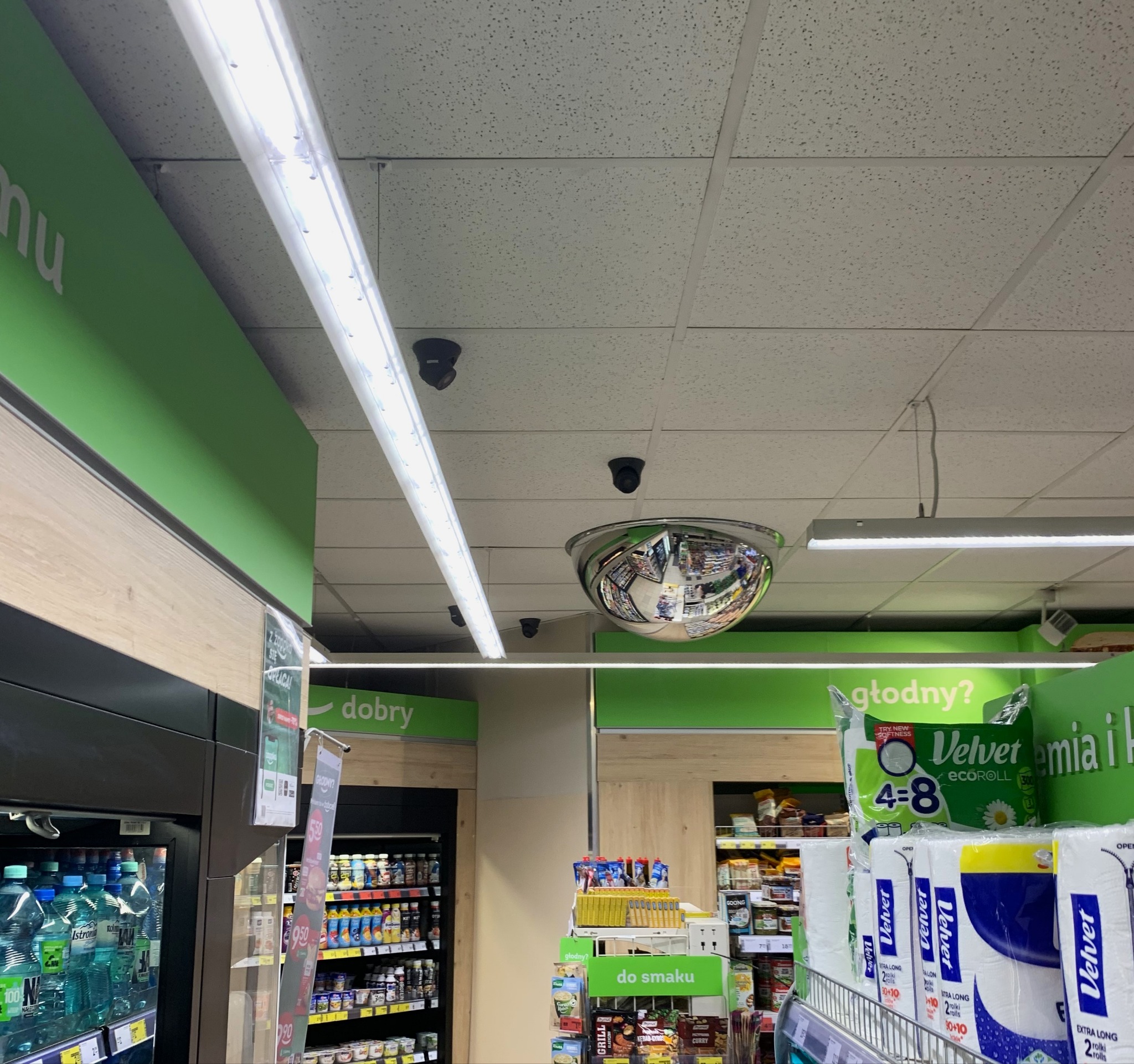 Widok na sufit sklepu z zainstalowanymi kamerami monitoringu, oświetleniem liniowym i półkulistym lustrem odblaskowym, odbijającym alejki sklepowe.