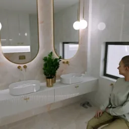 Elegancka łazienka z marmurowymi ścianami, podwójną umywalką nablatową, złotymi detalami, rośliną w złotej doniczce i dużym oknem, z siedzącą kobietą w tle.