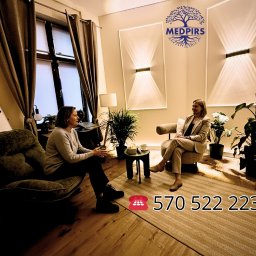 MedPirs Psychologia Psychoterapia - Dwie kobiety w jasnym gabinecie podczas rozmowy, otoczone roślinami. Na ścianie dekoracyjne lampy i logo Medpirs. W tle okno z zasłonami i biurko.