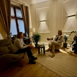 Dwie kobiety w jasnym gabinecie podczas rozmowy. Minimalistyczny wystrój, stonowane kolory, lampy ścienne z efektem świetlnym, rośliny doniczkowe, dywan w jodełkę.