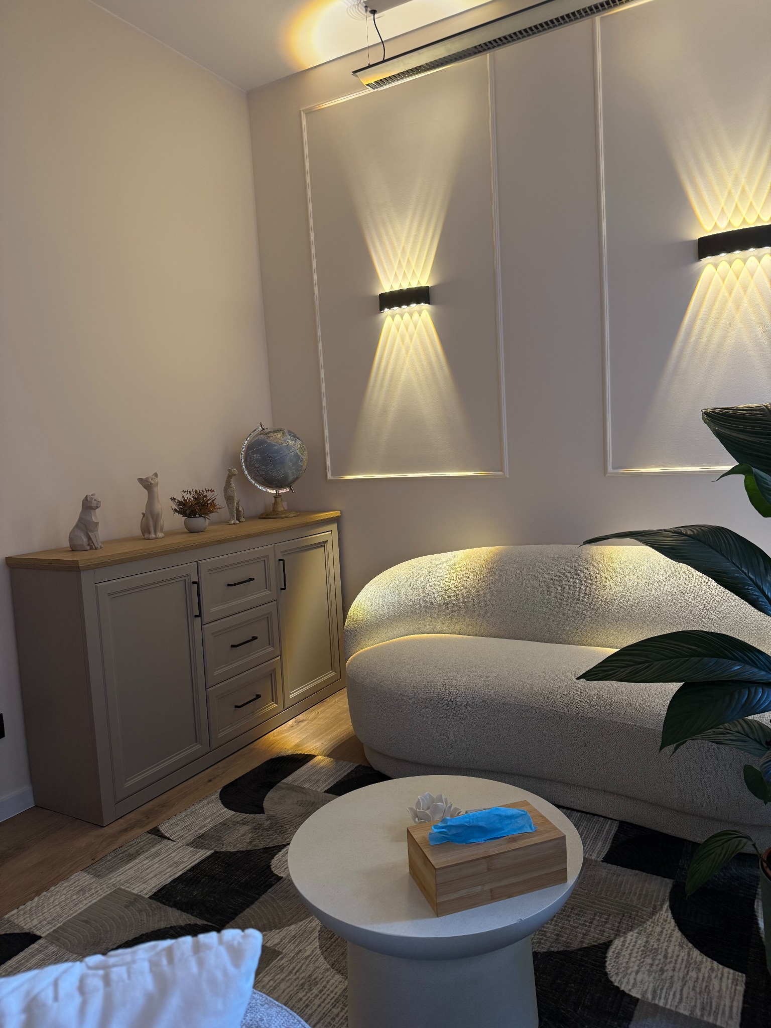 Jasne wnętrze gabinetu z sofą, stolikiem i komodą. Na stoliku chusteczki higieniczne. Na ścianie dekoracyjne panele z oświetleniem. Przytulna aranżacja.