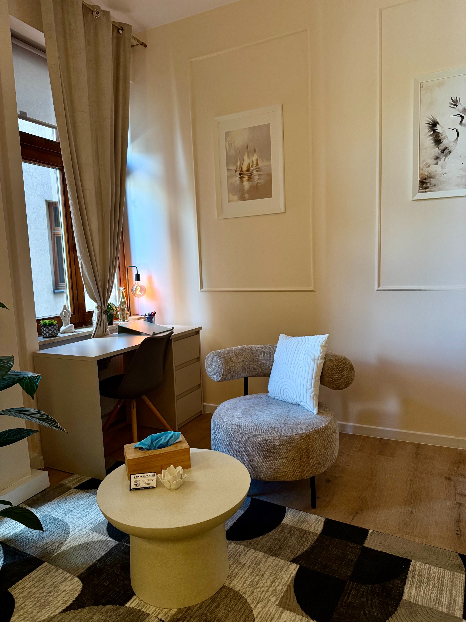 Jasny gabinet z biurkiem, okrągłym fotelem i stolikiem kawowym. Na ścianach obrazy w białych ramach. Przytulne i profesjonalne wnętrze.