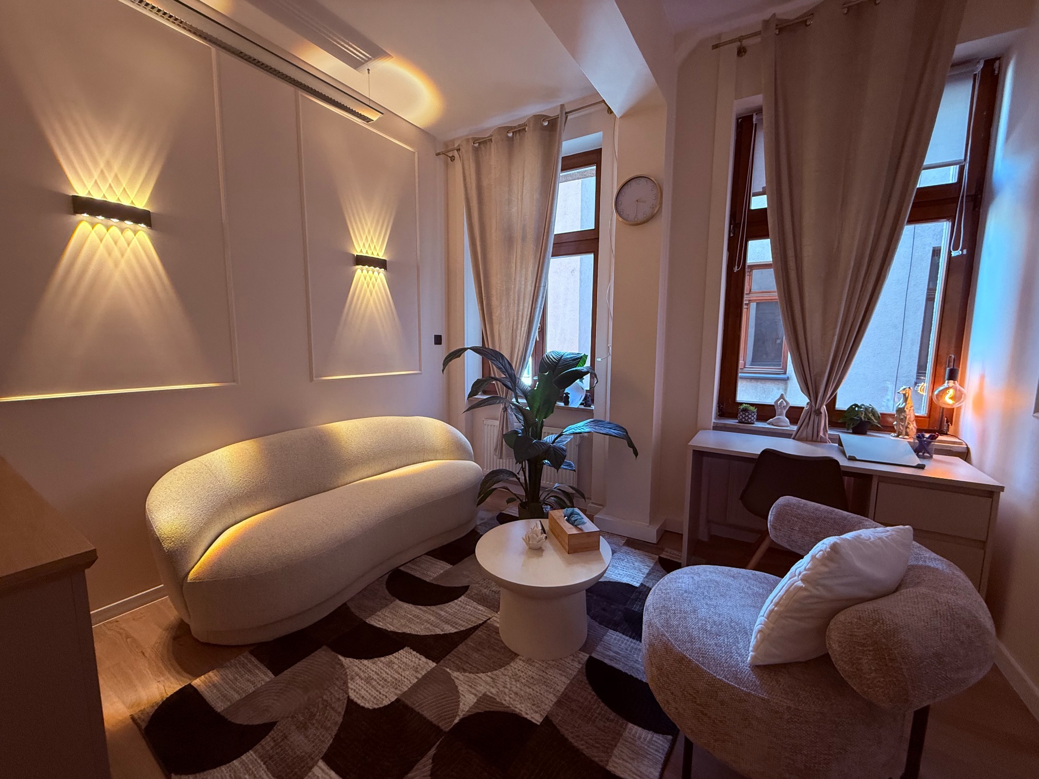 Przytulny gabinet z sofą, fotelem i stolikiem kawowym, oświetlony lampami ściennymi. Na biurku przy oknie widoczne dekoracje i lampka.