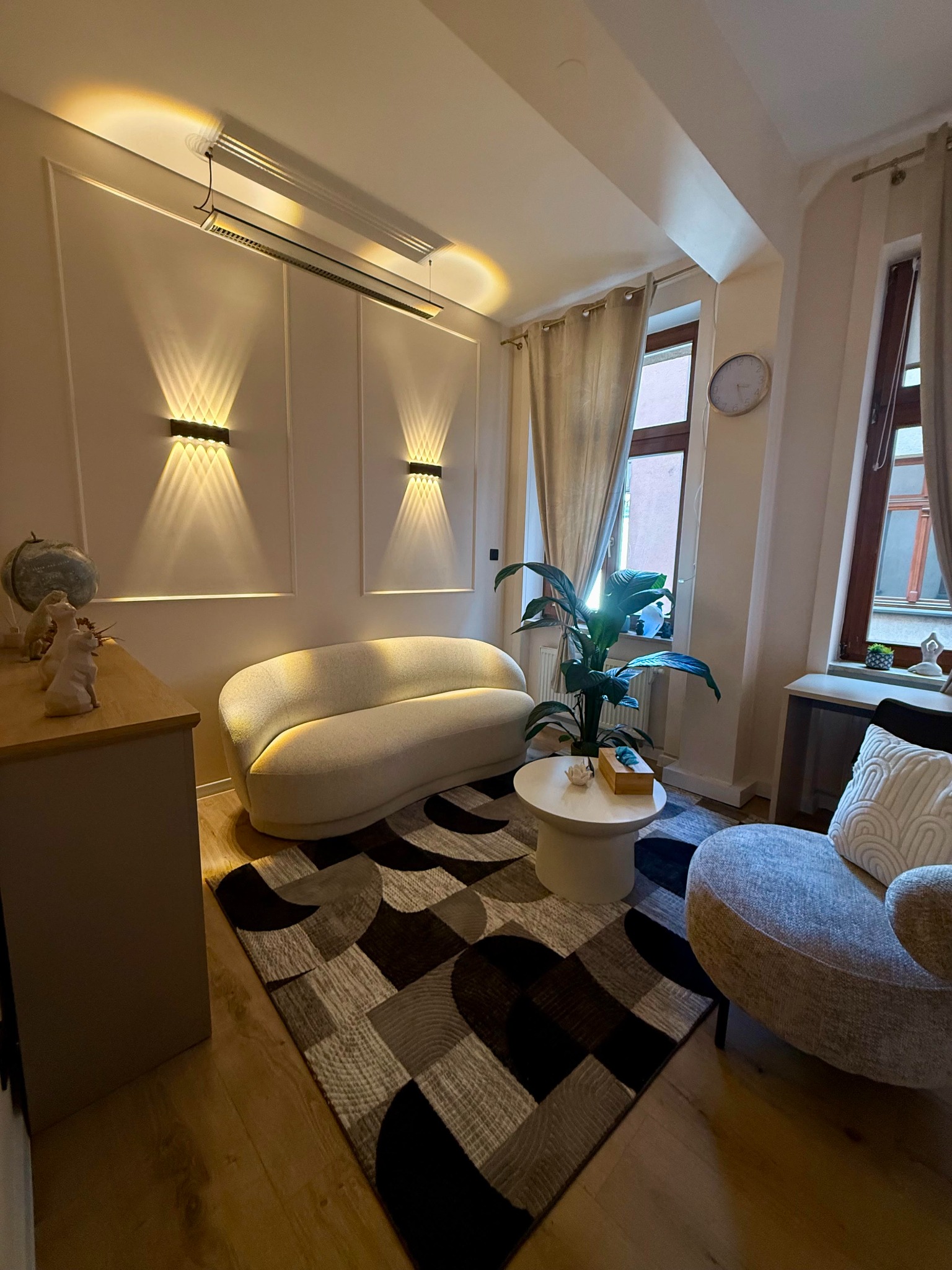 Przytulny gabinet z beżową sofą, wzorzystym dywanem i rośliną doniczkową na okrągłym stoliku. Dwa okna z zasłonami, oświetlenie ścienne i drewniana podłoga.