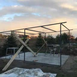 Konstrukcja stalowa altany w trakcie spawania, widoczne deski podpierające, stół spawalniczy i narzędzia na betonowej wylewce, w tle drzewa iglaste i zachodzące słońce.