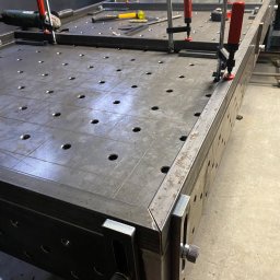 AP Welding Concept - Stół spawalniczy z otworami, narzędziami i uchwytami, w trakcie budowy, widoczne elementy mocujące i szlifierka kątowa.