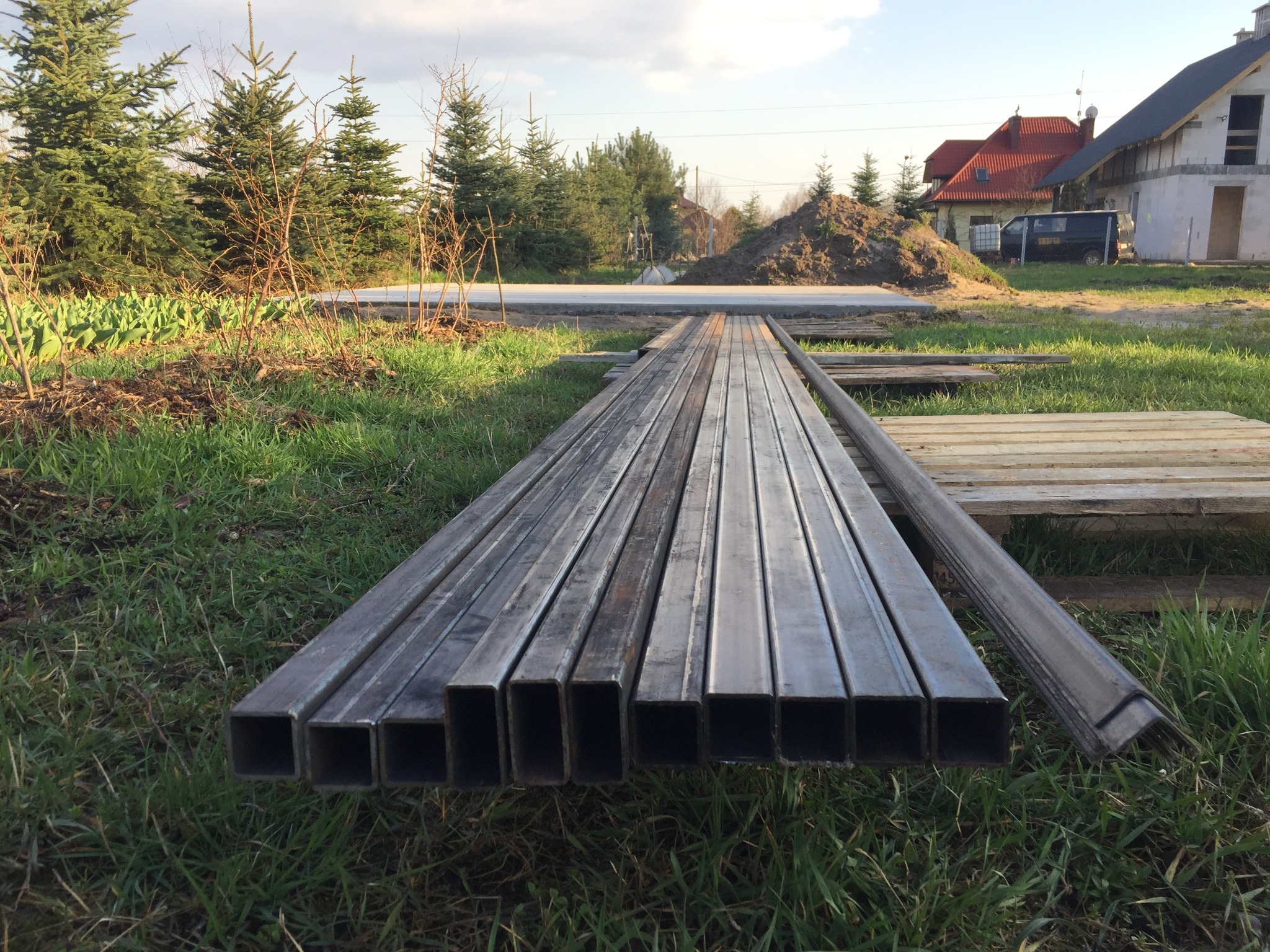 Stalowe profile kwadratowe ułożone na trawie w pobliżu placu budowy.