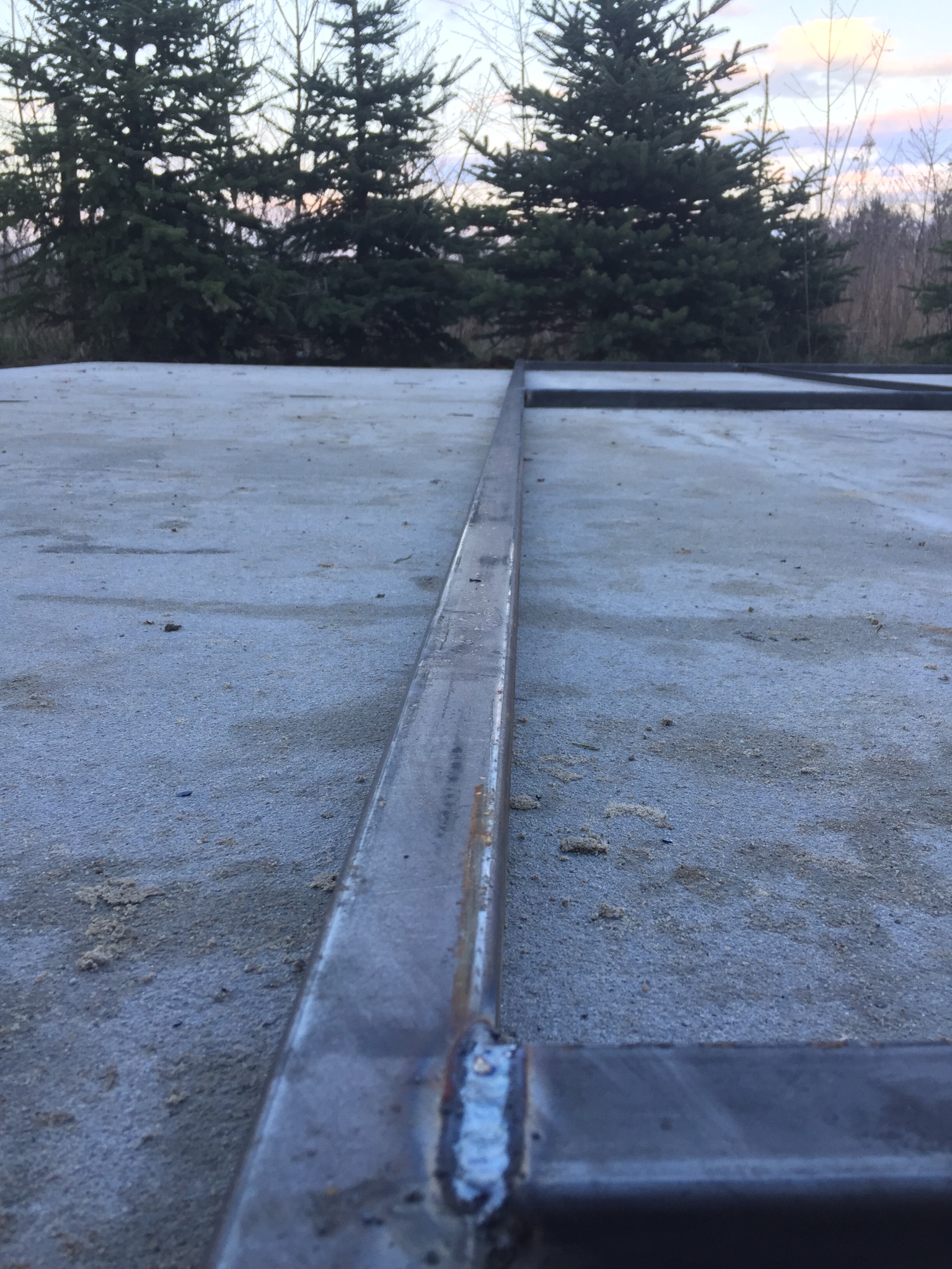 Metalowa konstrukcja spawana, prawdopodobnie podstawa balkonu lub tarasu, widoczna na tle betonowej powierzchni i drzew iglastych.