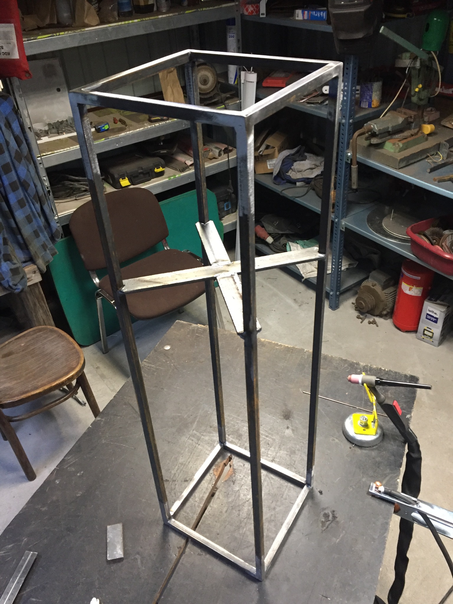 Metalowa konstrukcja w kształcie prostopadłościanu, w trakcie spawania w warsztacie, widoczny uchwyt spawalniczy i narzędzia na stole roboczym.