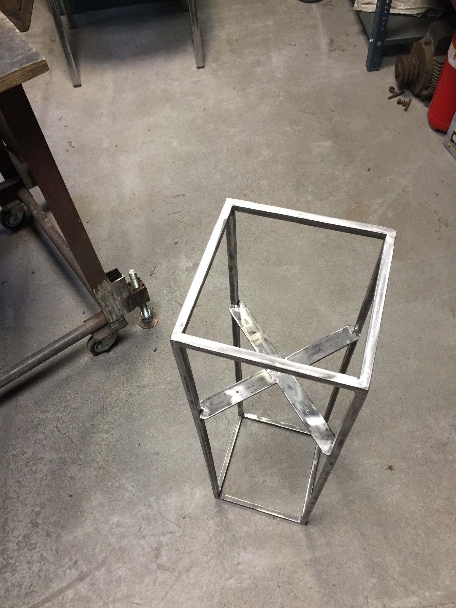 Surowa, metalowa konstrukcja w kształcie sześcianu z wewnętrznym wzmocnieniem w postaci krzyżujących się płaskowników, widoczna na szarym betonowym podłożu w warsztacie.