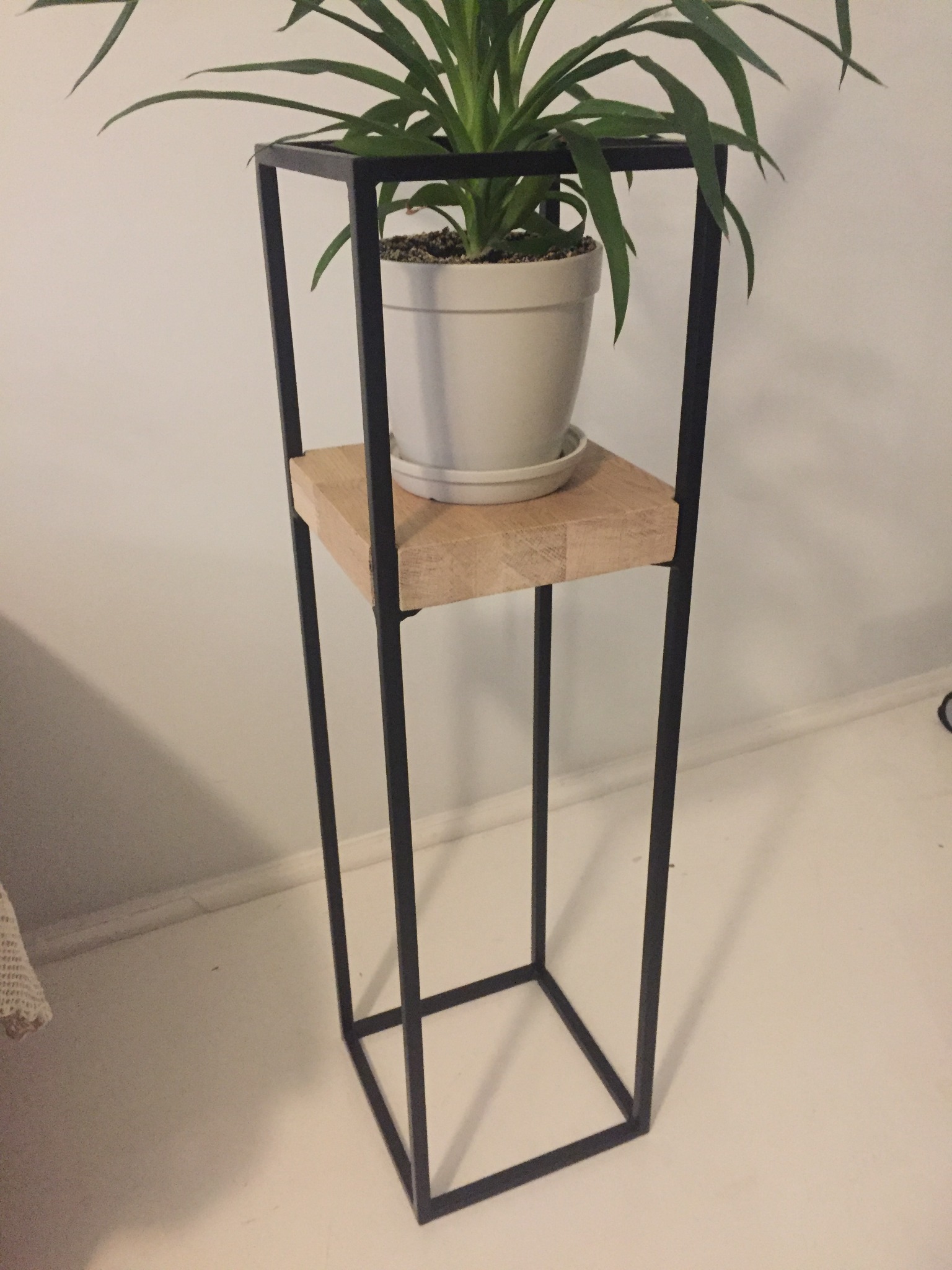 Czarny, spawany kwietnik z pręta kwadratowego, z dębowym olejowanym blatem, na którym stoi biała doniczka z zieloną rośliną.