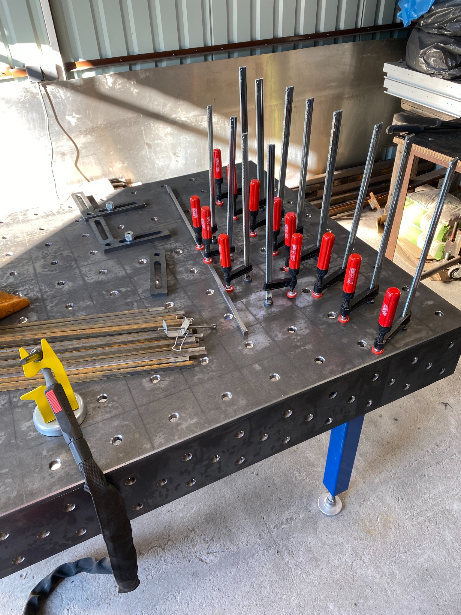 Stół spawalniczy z zamocowanymi elementami metalowymi za pomocą ścisków, widoczny uchwyt spawalniczy i przygotowane pręty do spawania.