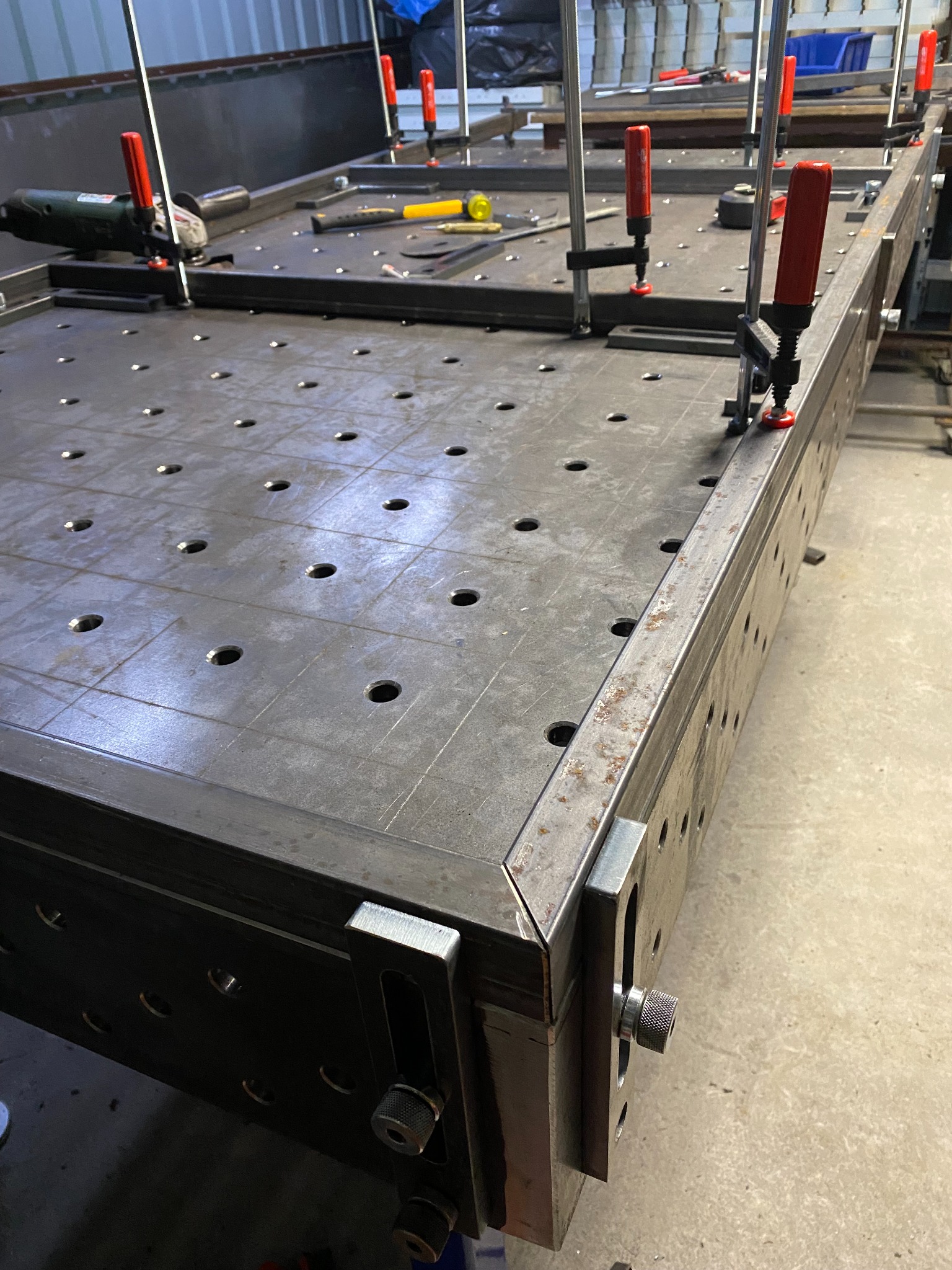 Stół spawalniczy z otworami, narzędziami i uchwytami, w trakcie budowy, widoczne elementy mocujące i szlifierka kątowa.