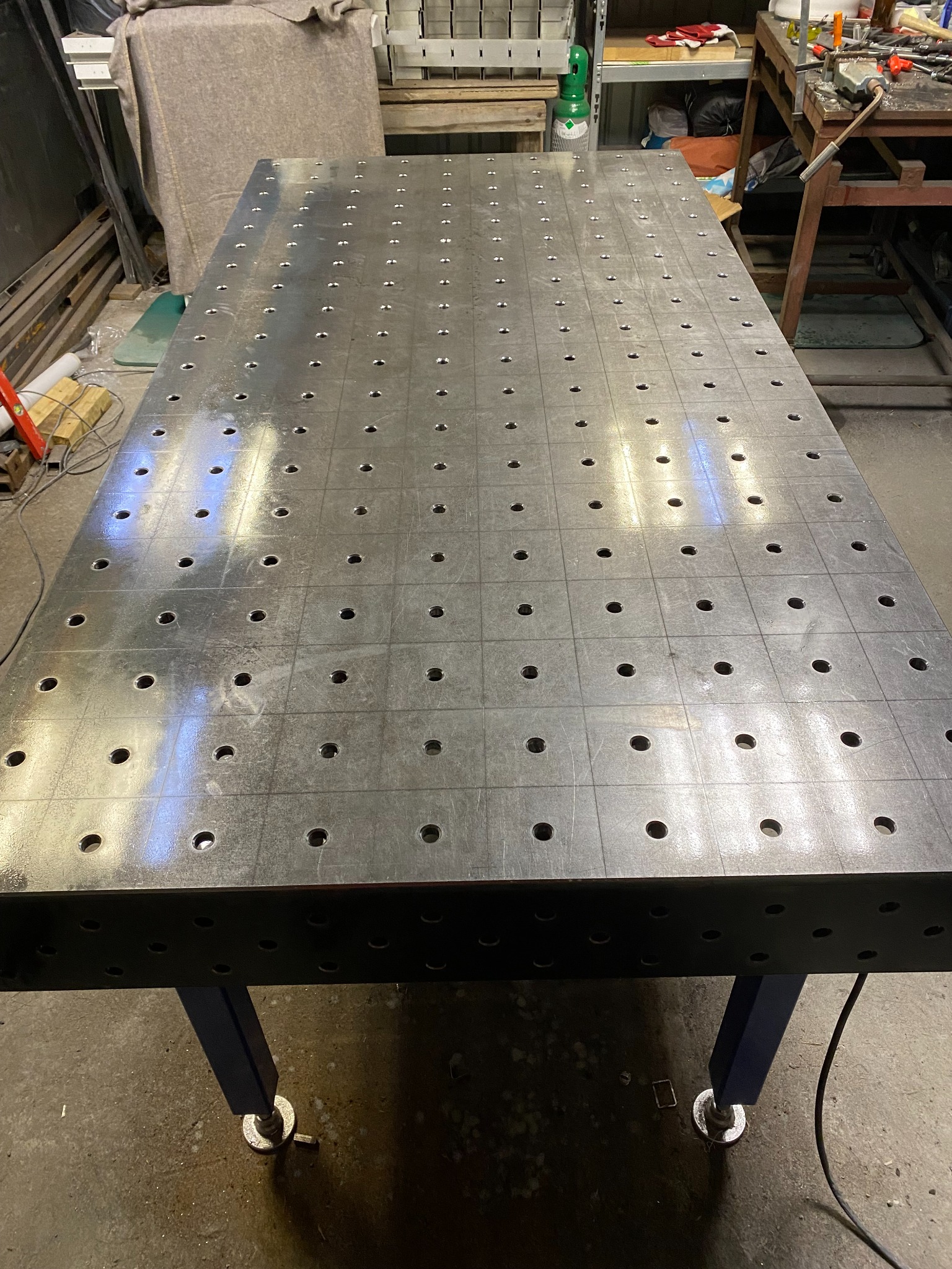 Stół spawalniczy z perforowanym blatem, ustawiony w warsztacie, widoczne ślady użytkowania i regulowane nóżki.